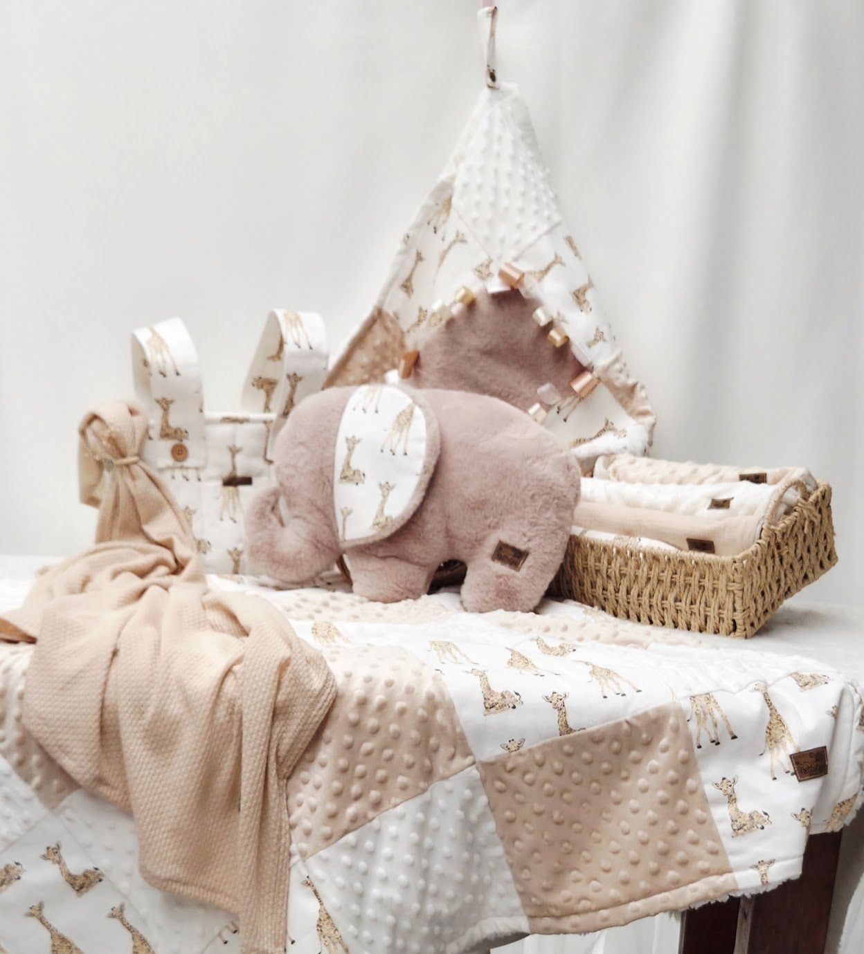 Neutral-toned baby nursery set with plush elephant, giraffe-print blanket, wicker basket, and teepee decor.