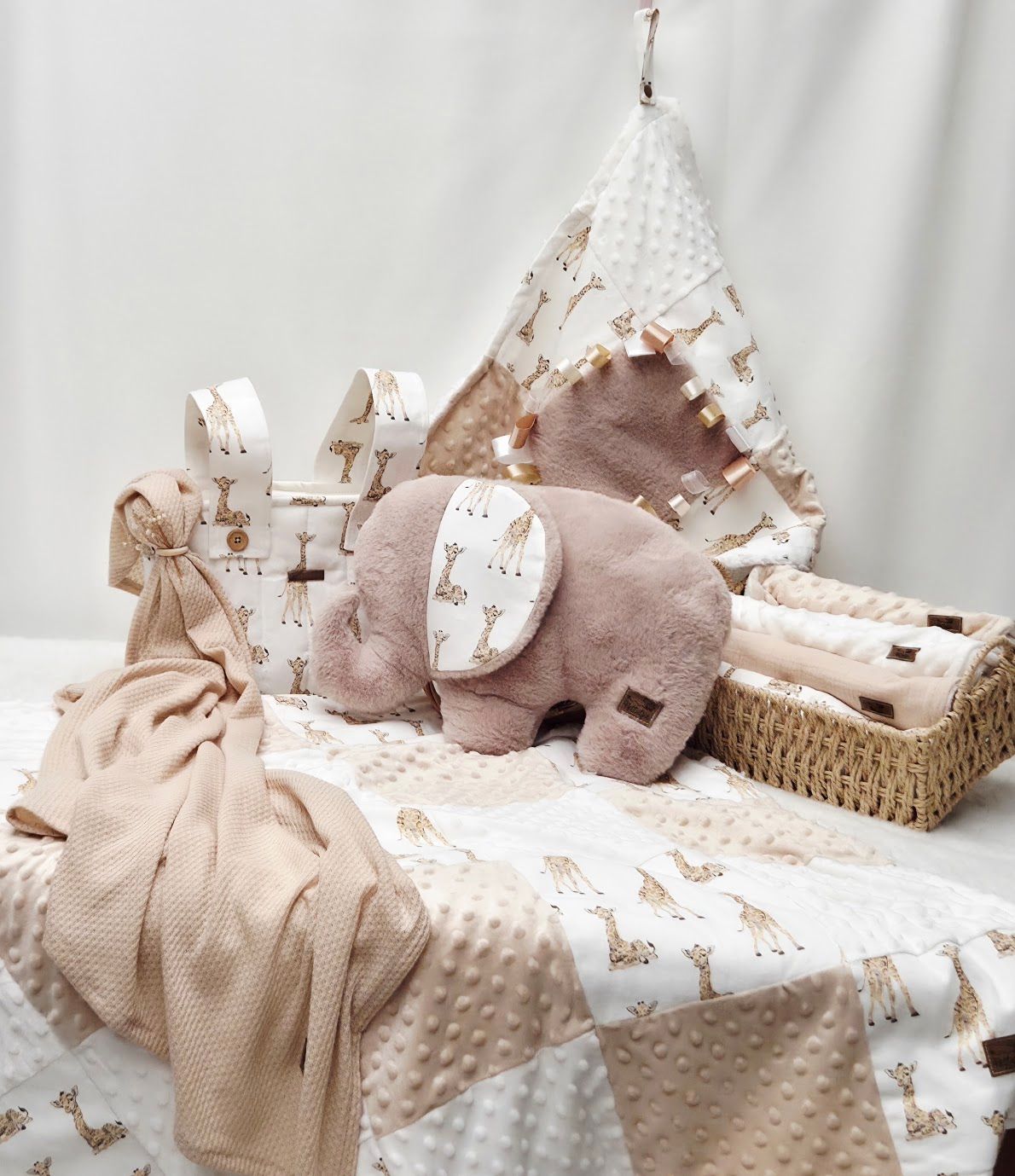 Soft baby nursery set with elephant plush, giraffe print teepee, minky blanket, and wicker basket in blush tones.