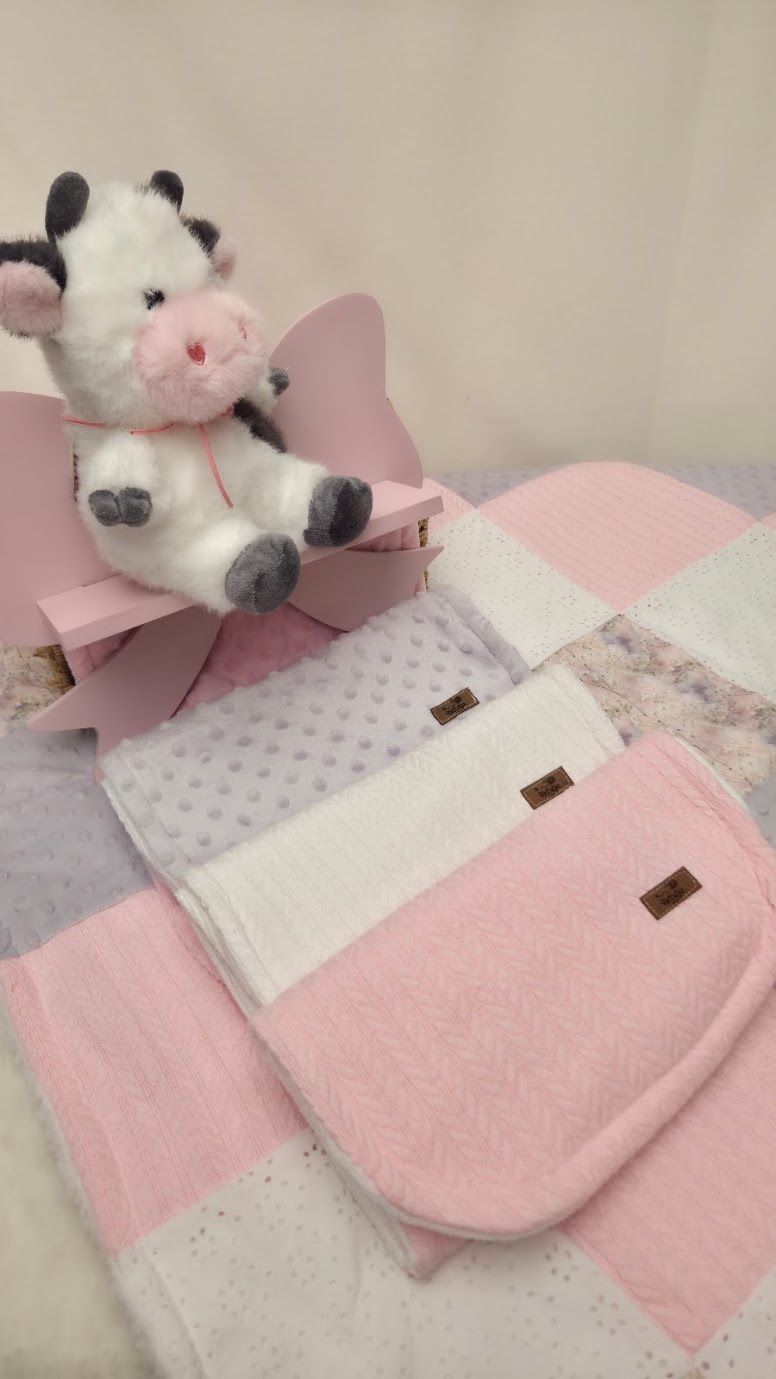 Pink baby gift set with cow plush toy, quilted blanket, and soft burp cloths on a pink checkered background.