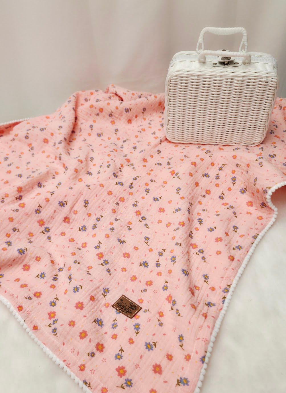 Pink floral muslin baby blanket with white pompom trim displayed next to a white wicker basket purse.