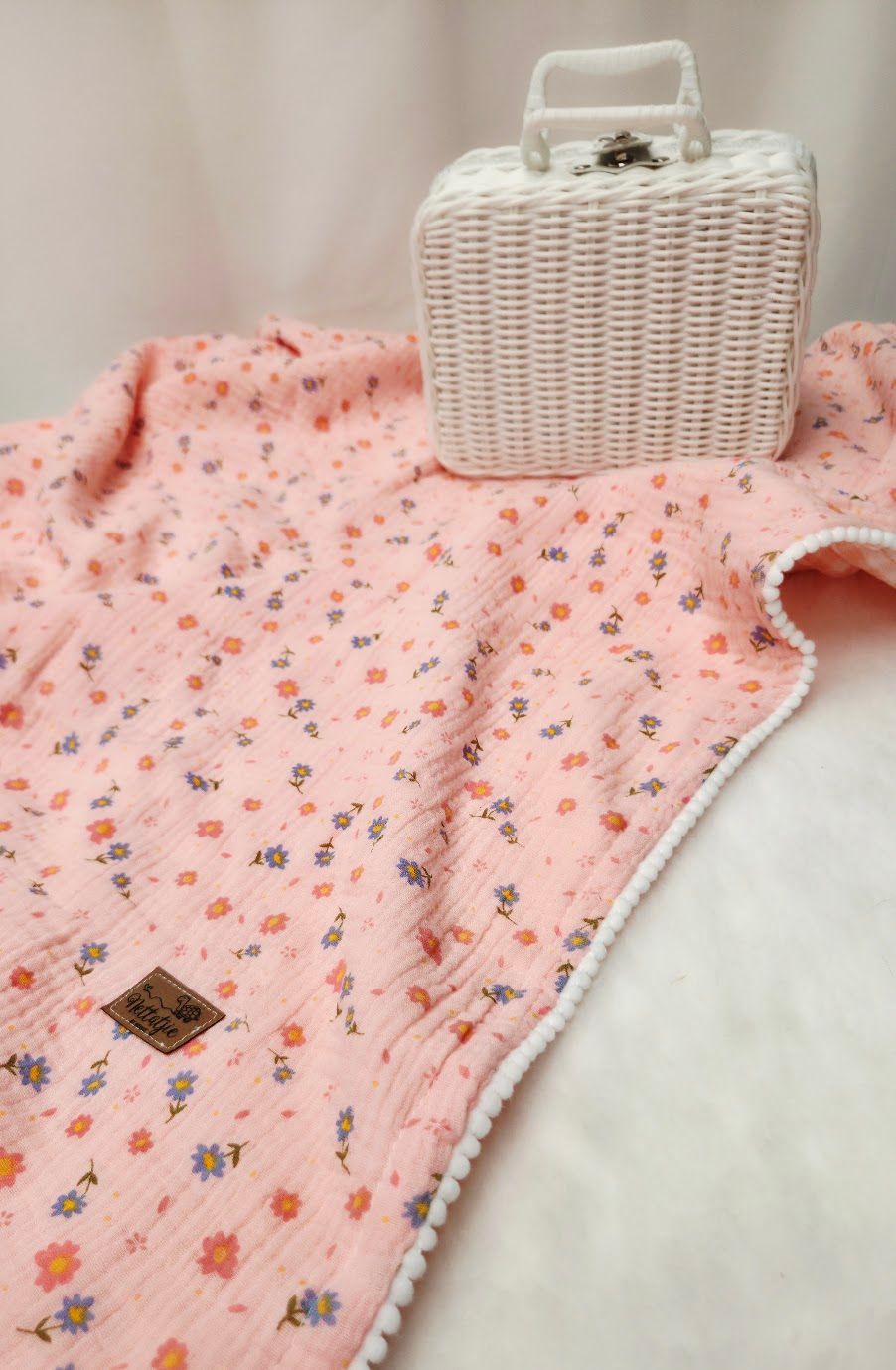 Pink floral muslin baby blanket with pom-pom trim laid flat beside a white wicker suitcase.
