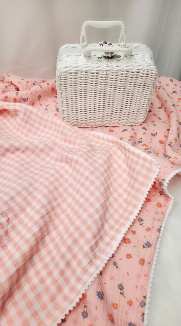 Pink gingham and floral reversible baby blanket with pom-pom trim, displayed with a white wicker suitcase.