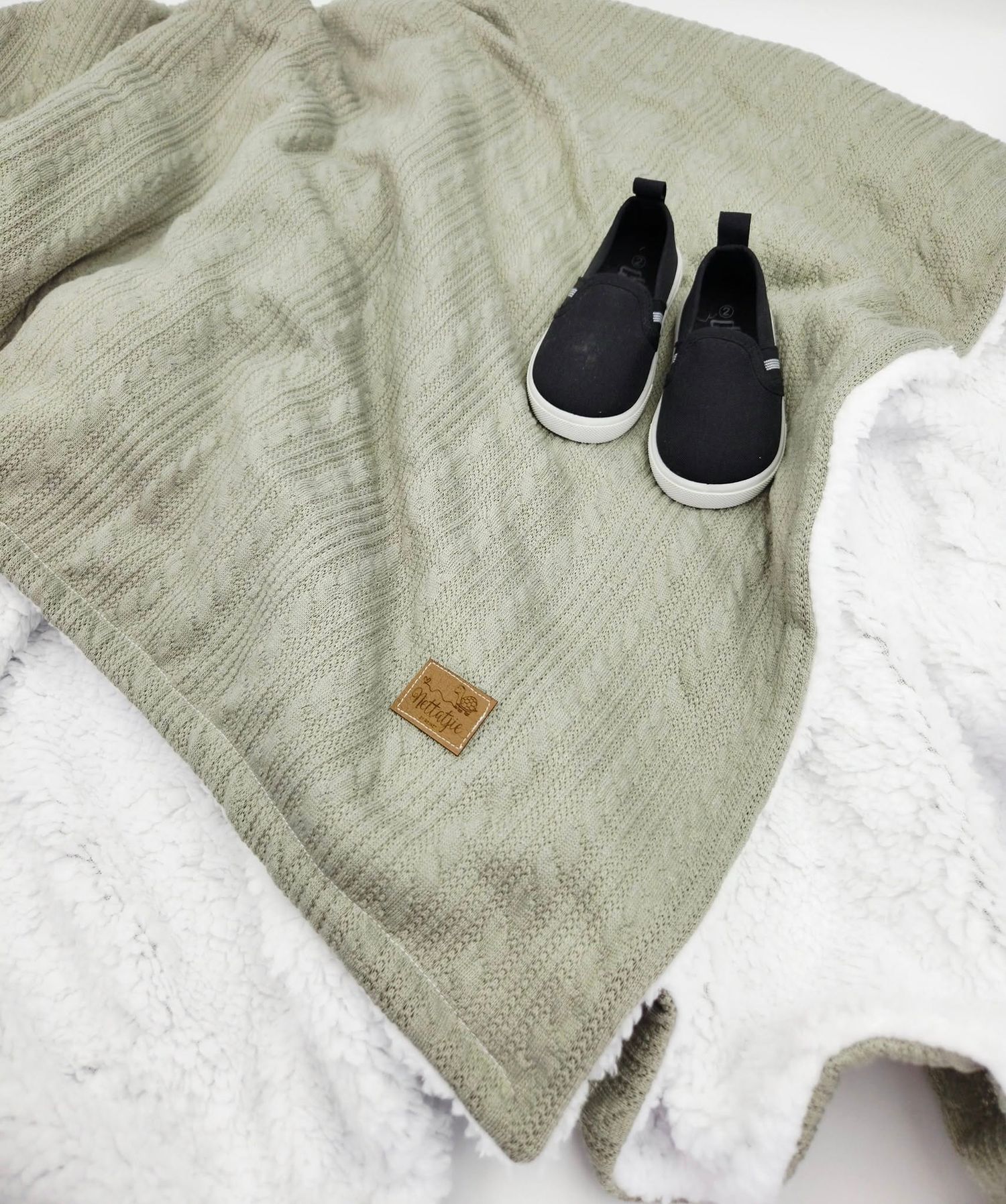 Sage green knitted baby blanket with white trim and black baby shoes.
