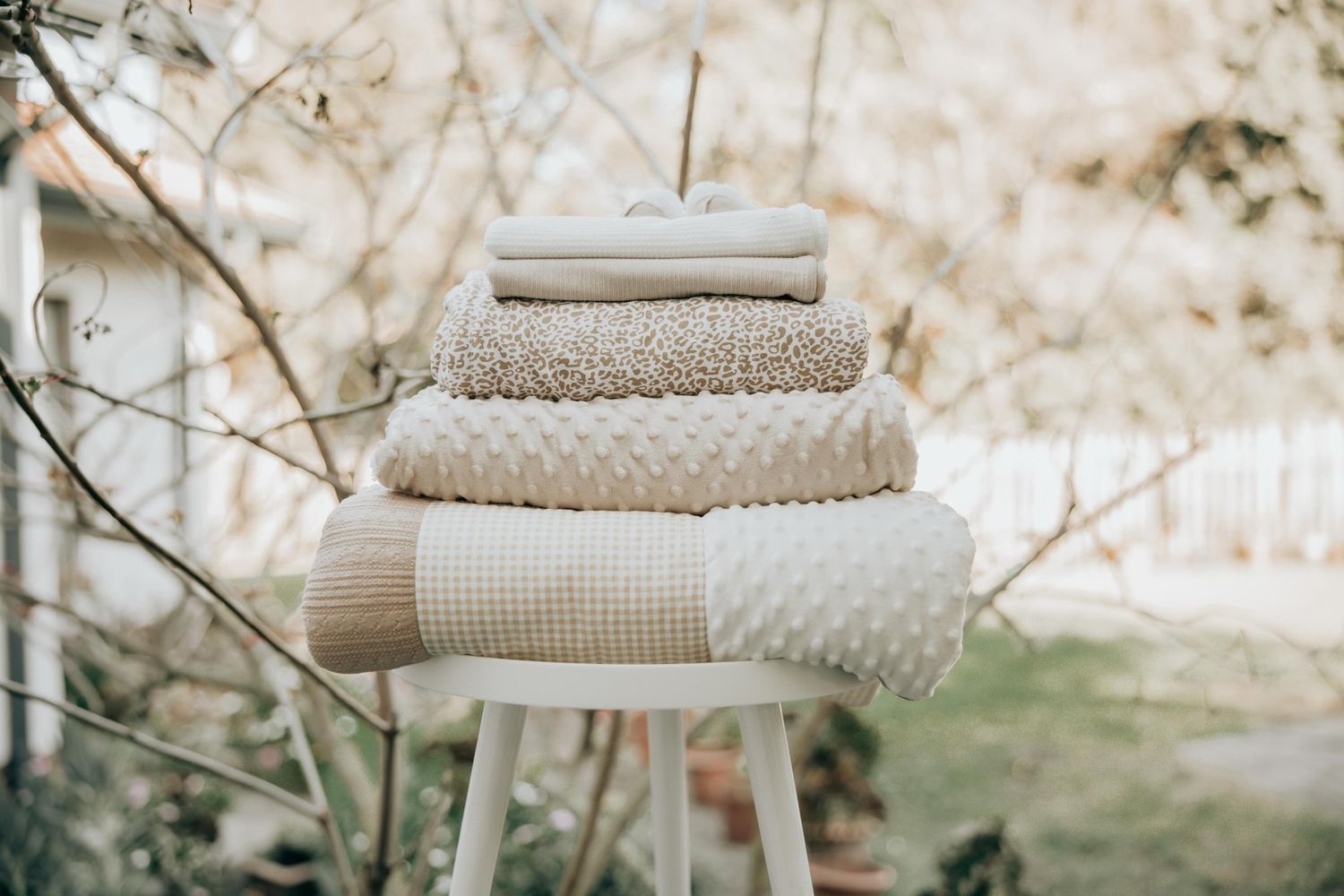 Soft neutral knit blankets and textiles stacked in cozy layers on white furniture in natural lighting.