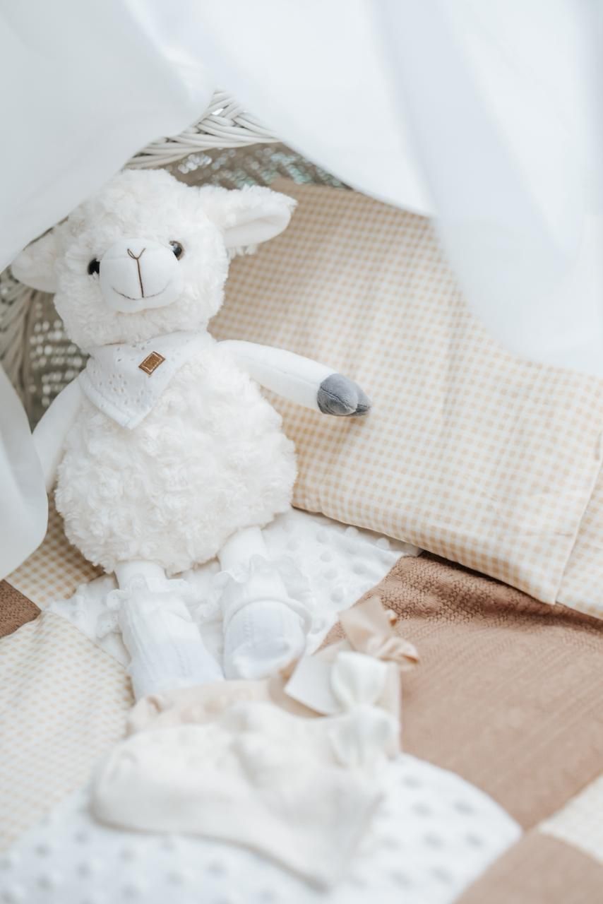 White plush lamb toy on beige checkered bedding with baby clothes and accessories in soft neutral tones.