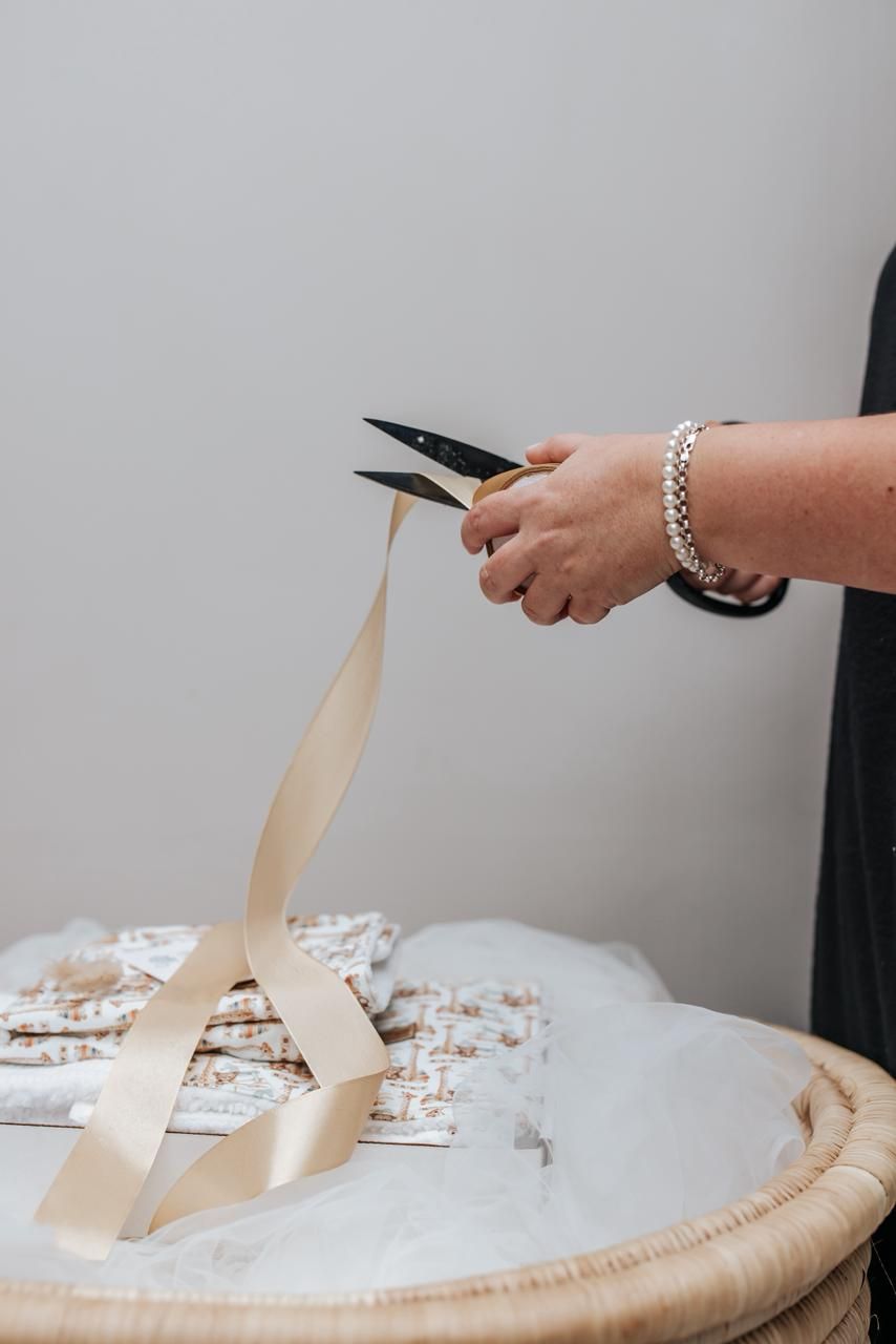 Hands using scissors to cut strips of light colored paper or fabric on a white surface.
