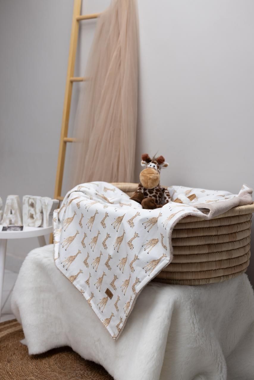 A cozy nursery corner with a rope basket, giraffe print blanket, wooden ladder, and plush giraffe toy on white linens.
