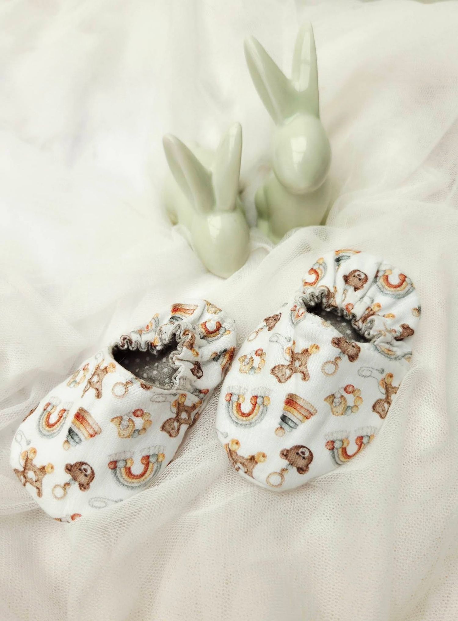 Handmade baby shoes with colorful rainbow and animal print pattern displayed beside ceramic bunny figurines on white fabric.