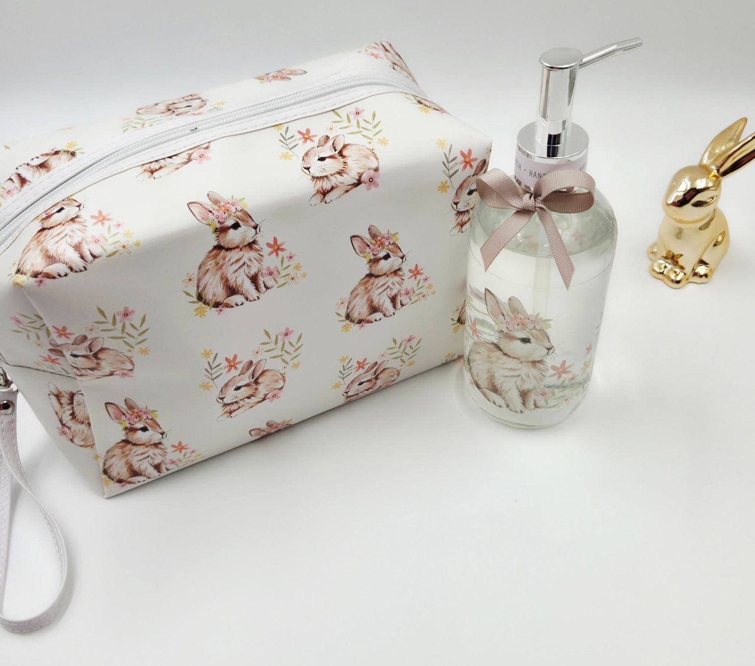 White cosmetic bag with bunny print pattern beside a glass soap dispenser and gold rabbit figurine.