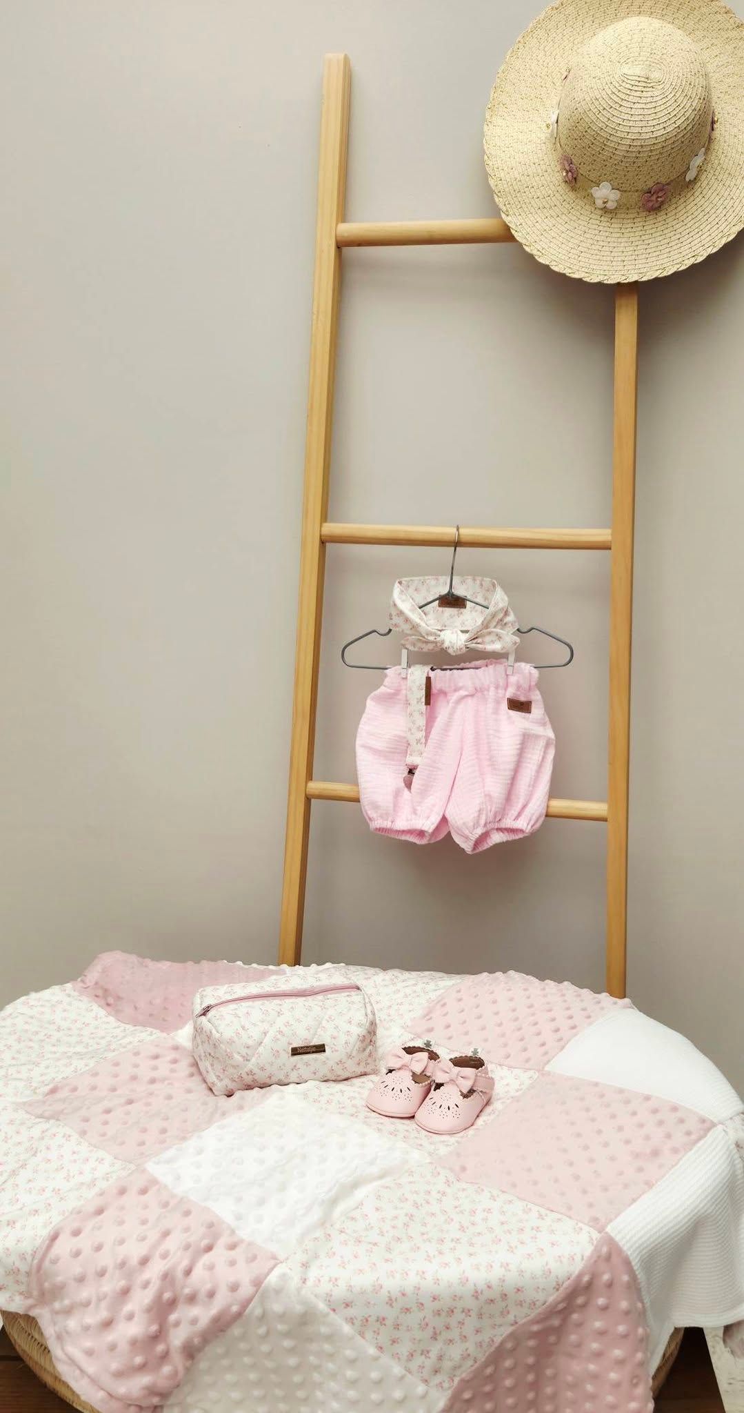 Pink baby girl set with romper, blanket, shoes, cosmetic bag, and straw hat displayed on wooden ladder.