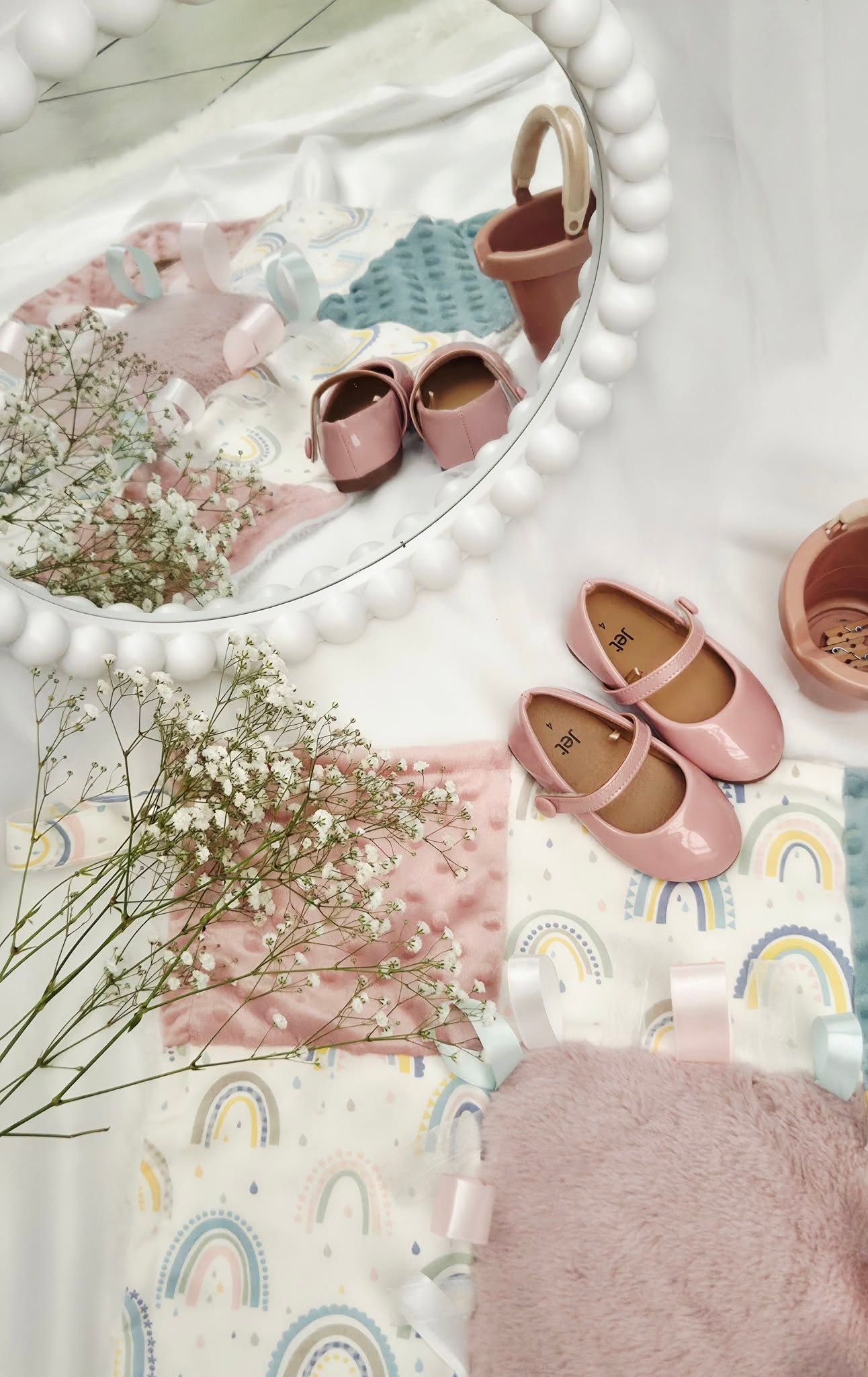 Pink baby Mary Jane shoes displayed with rainbow onesies, baby's breath flowers, and a round beaded mirror.