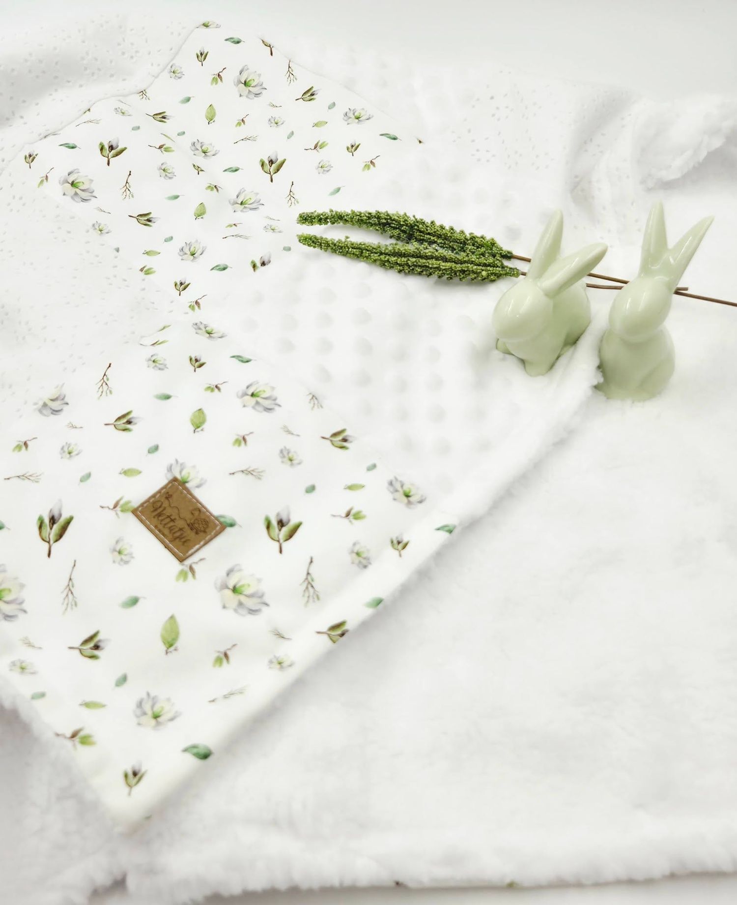 White floral baby blanket with minky dot fabric, small leather label, green sprig, and ceramic bunny decorations.