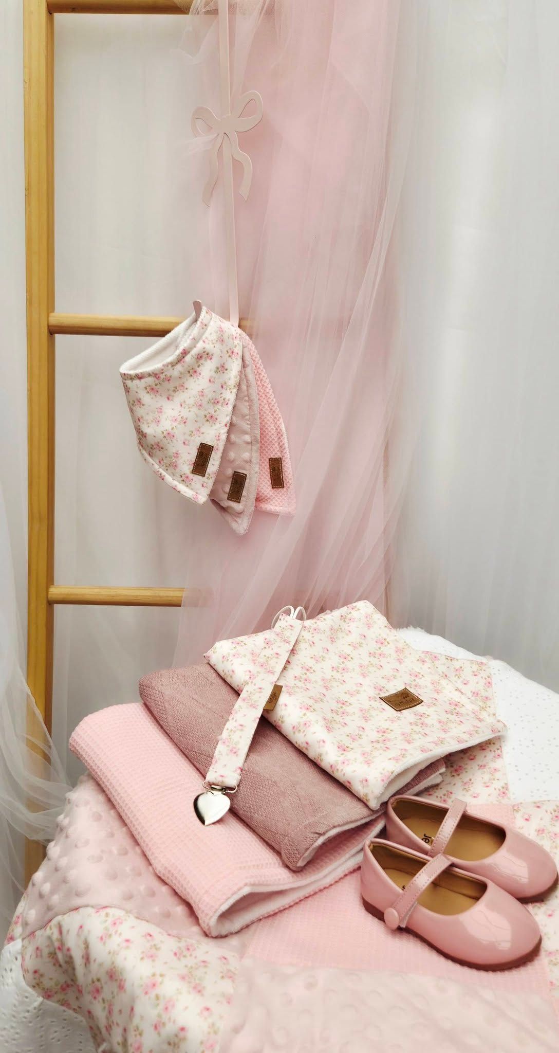 Pink baby girl accessories set with floral bag, bibs, ballet shoes, and blanket displayed on a ladder and bed.