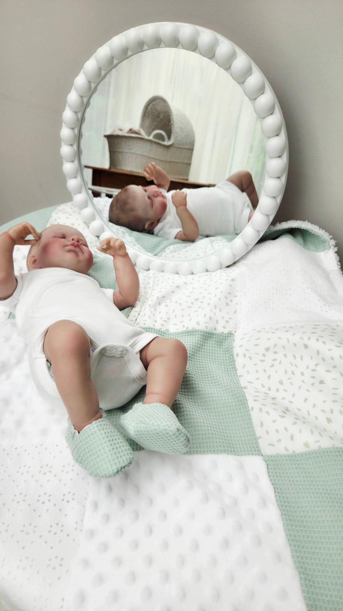 Newborn baby in white onesie and mint booties lying on patchwork quilt beside round decorative mirror in nursery.