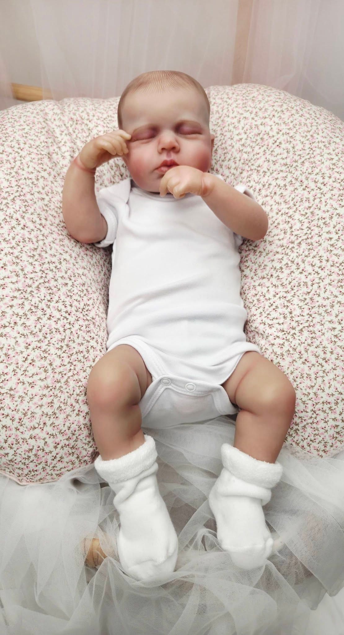 Realistic reborn baby doll in white onesie and socks, resting on a floral nursing pillow with eyes closed.