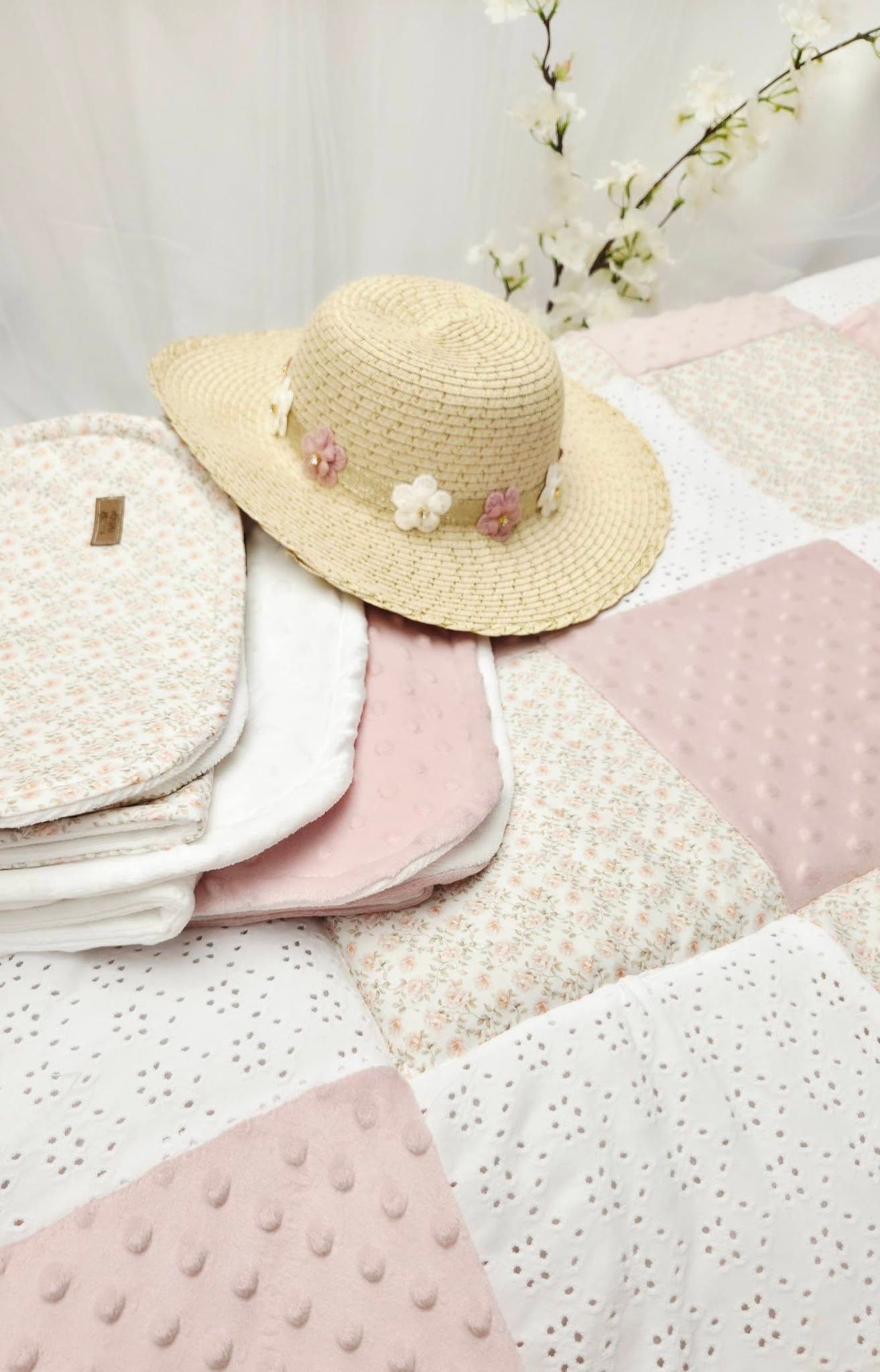Pastel patchwork quilt with pink and cream squares, straw hat with floral trim, and folded baby blankets on display.