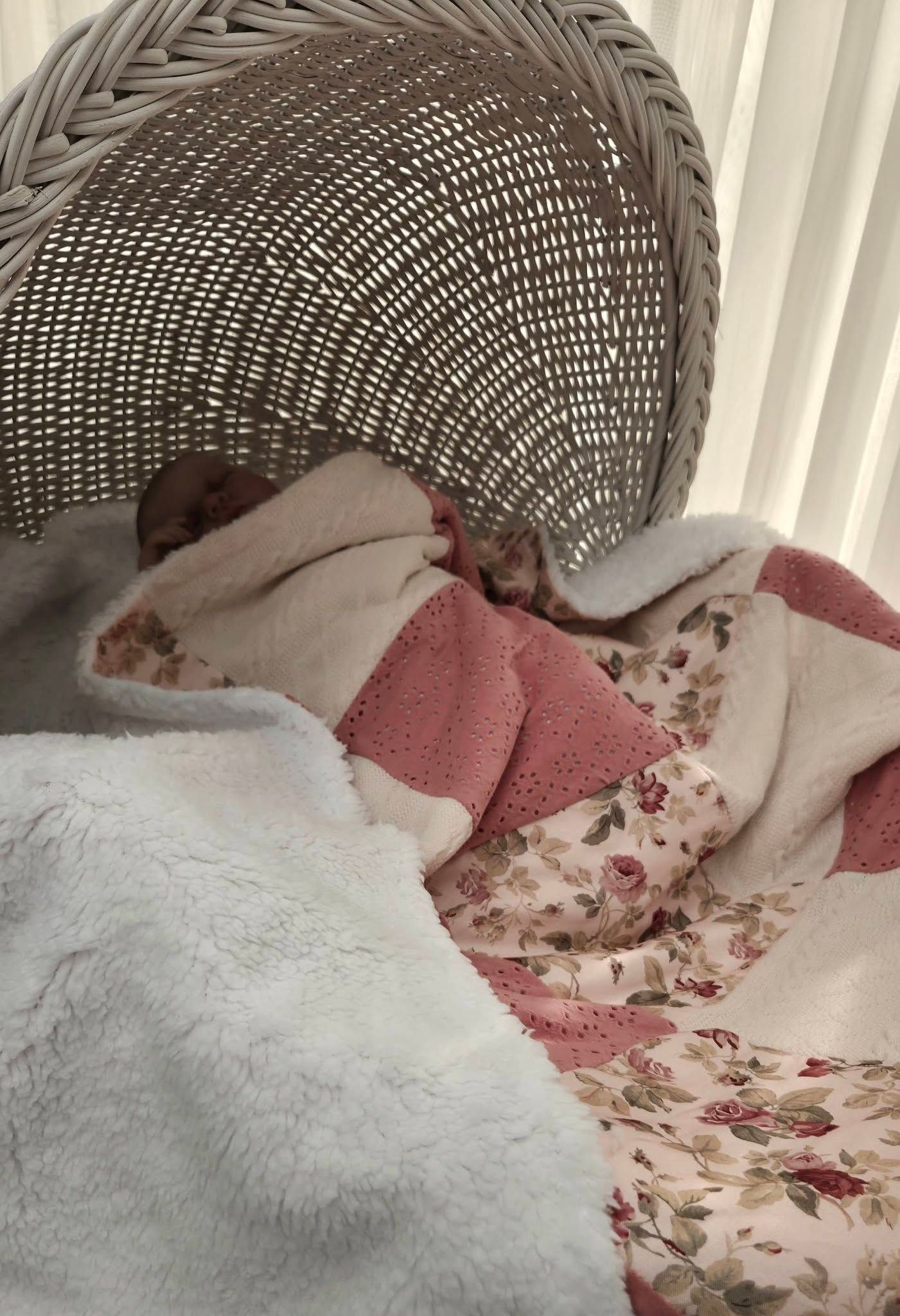 Newborn baby sleeping peacefully wrapped in pink floral and white blankets beside a wicker bassinet.