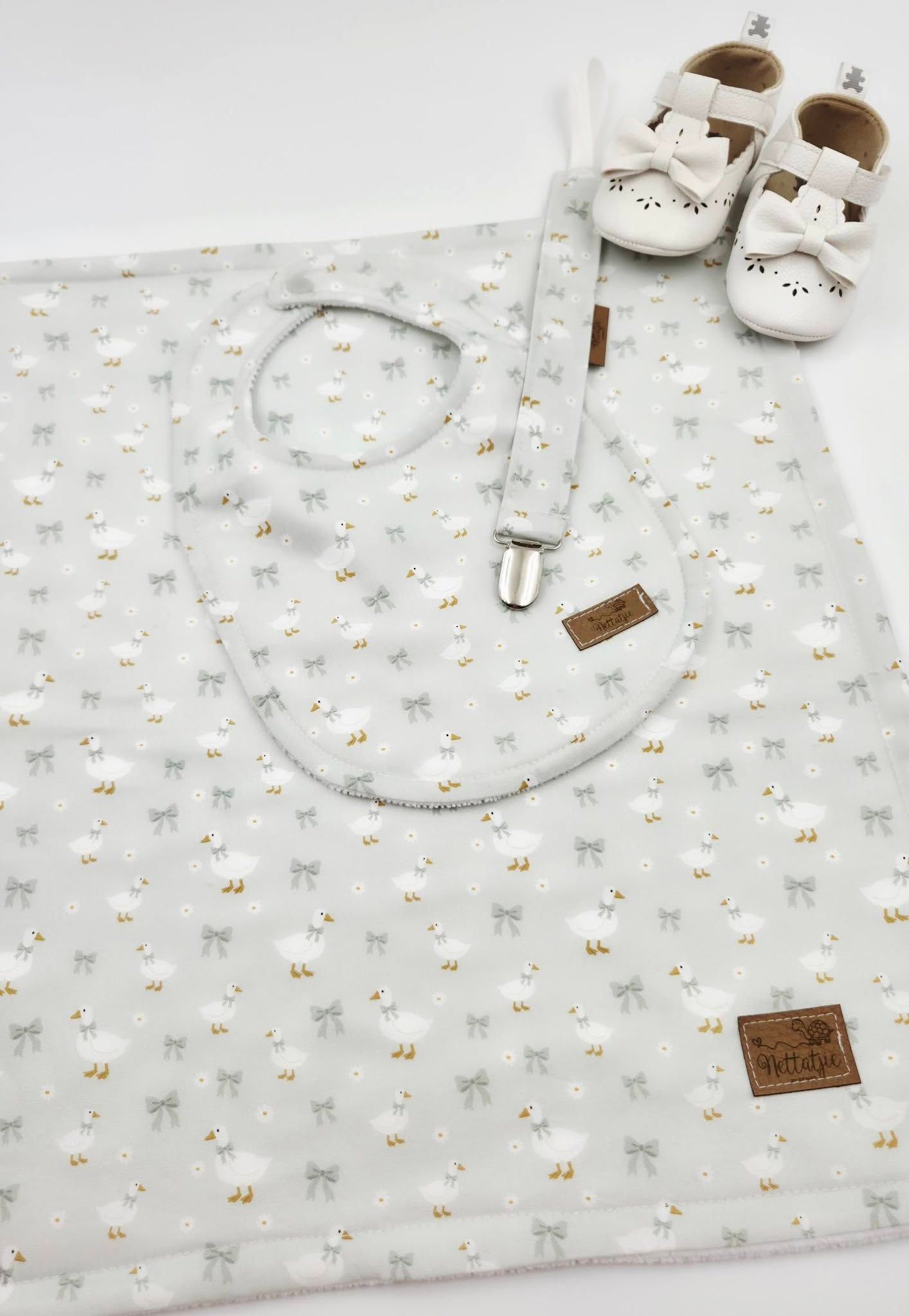 Baby gift set with sheep-print blanket, bib, pacifier clip, and white leather shoes on light background.