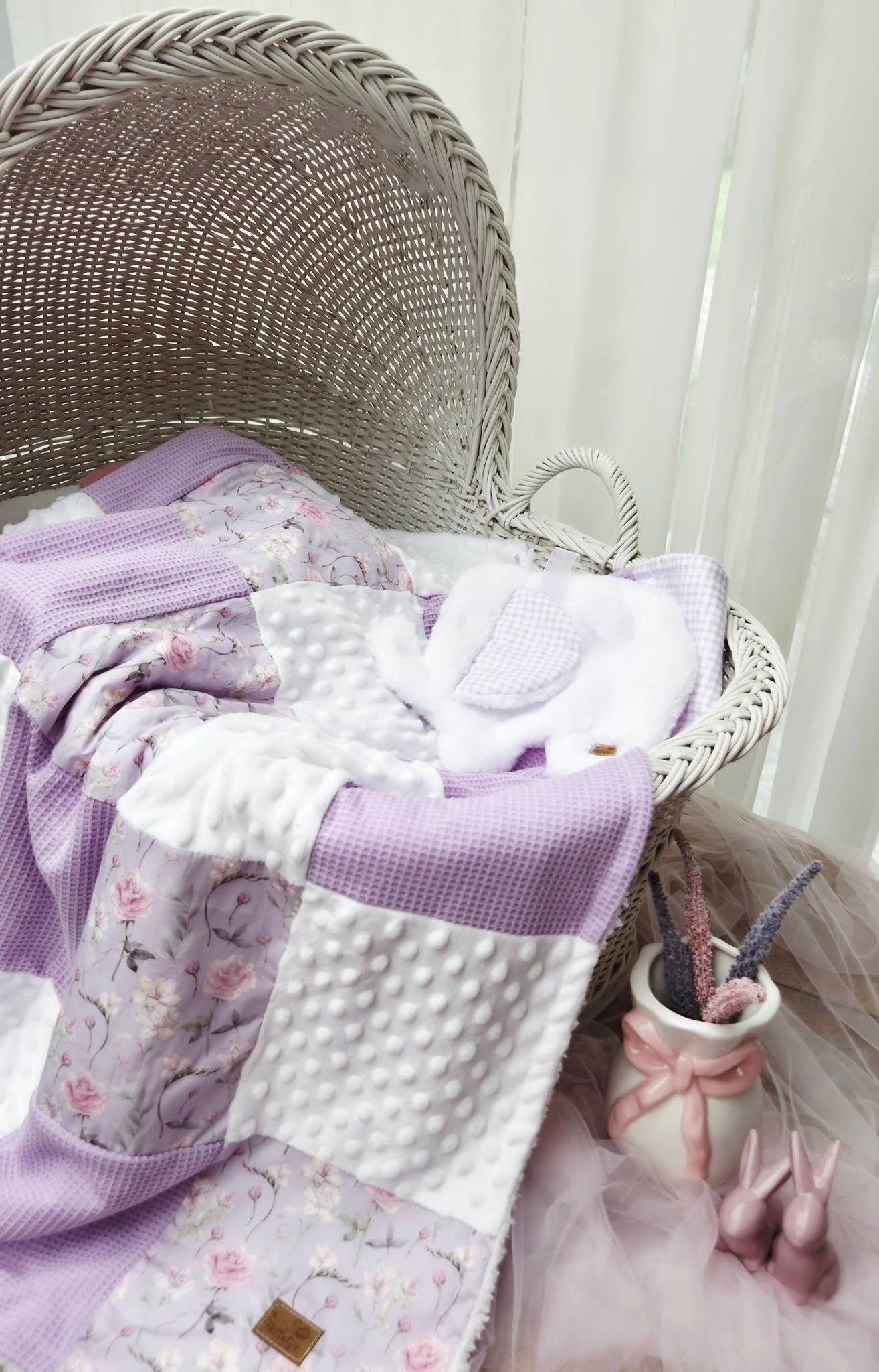 Lavender patchwork baby blanket with floral print draped in a wicker bassinet beside pink nursery decor.