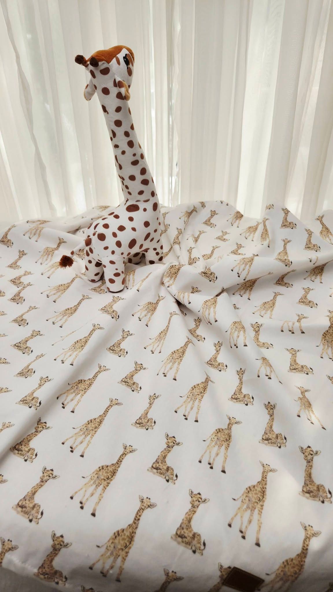 A stuffed giraffe toy sits upright on white fabric printed with a repeating pattern of illustrated giraffes.