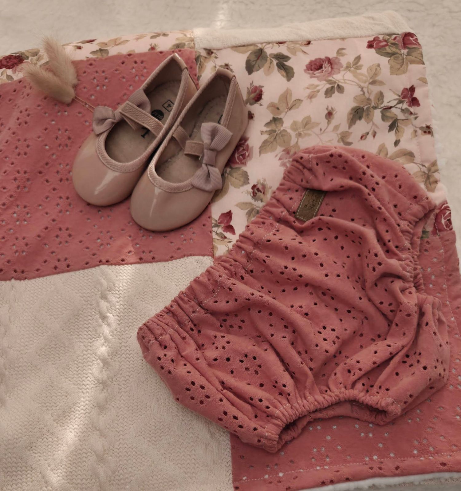 Pink baby girl outfit set with eyelet romper and matching bow Mary Jane shoes displayed on floral fabric.