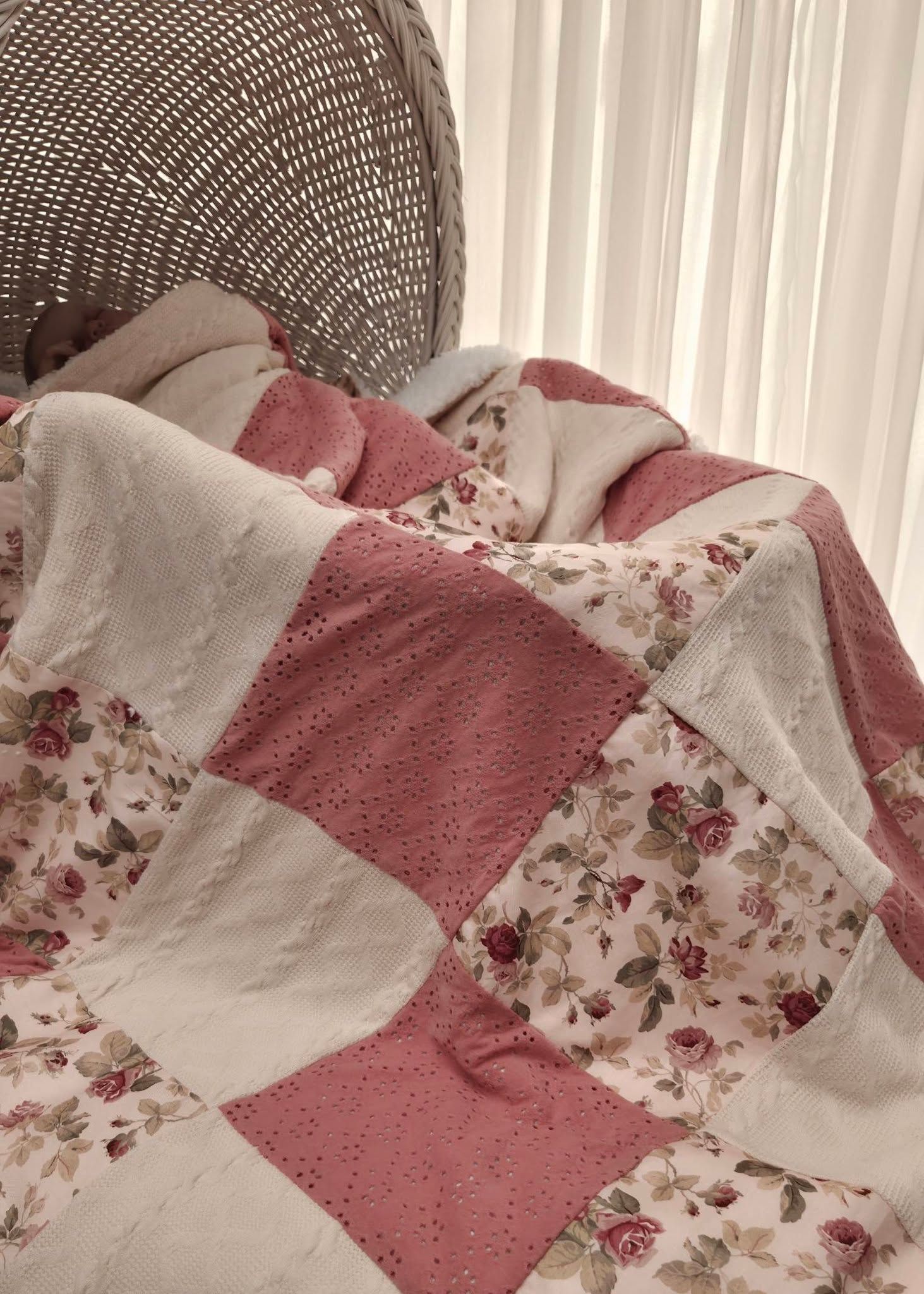 Pink and white patchwork quilt with floral rose print draped over a wicker chair near sheer curtains.