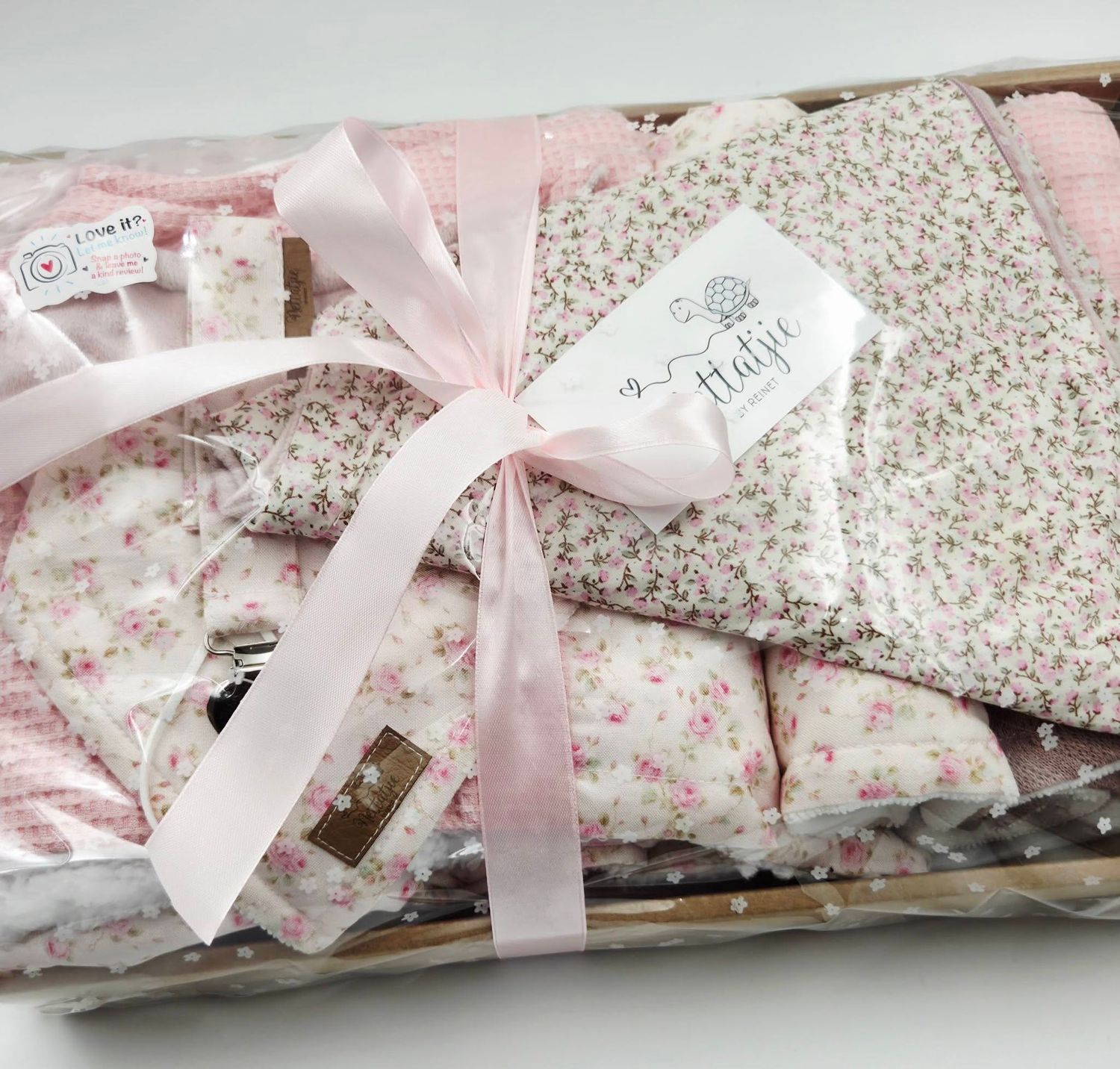 Pink floral baby gift set with ribbon bow, featuring soft blankets and accessories in a wicker basket.