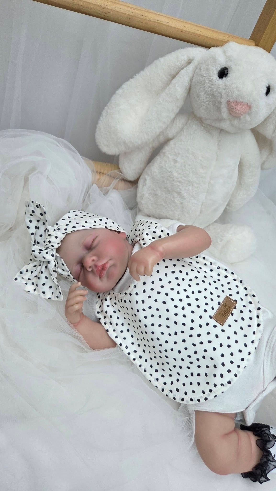 Realistic reborn baby doll in black and white heart print outfit with matching bow, posed beside a white plush bunny.