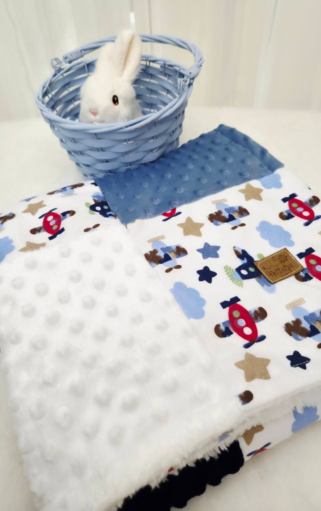 Baby blanket with airplane and star print, white minky dot fabric, blue textured side, and a white bunny toy in a blue basket.