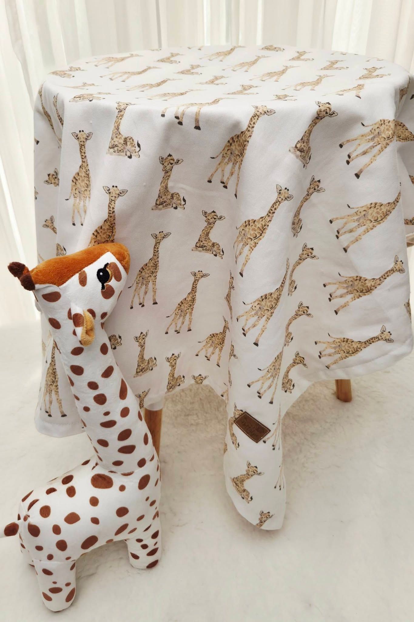 White tablecloth with giraffe print pattern displayed on a small table beside a plush giraffe stuffed animal toy.