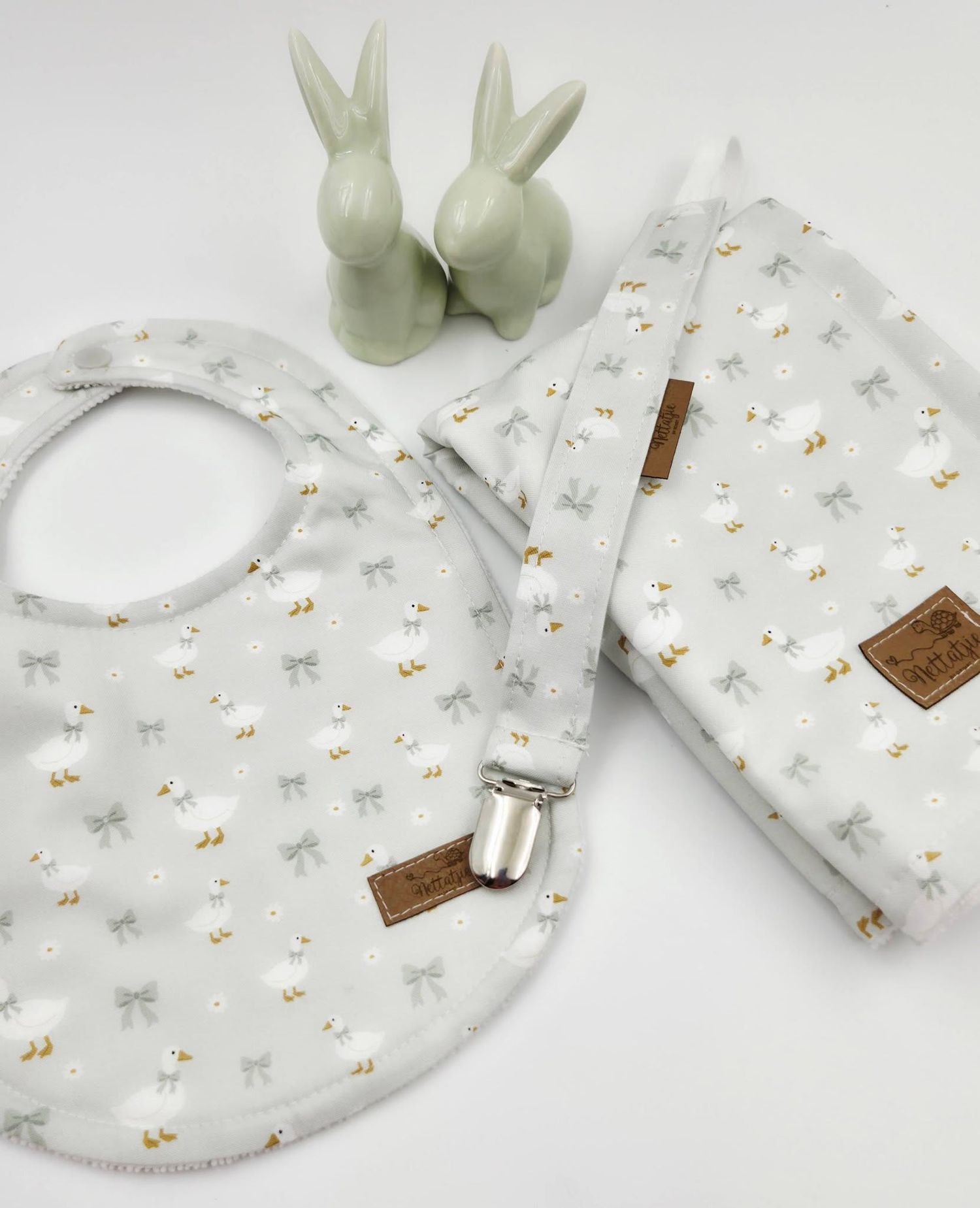 Gray floral baby bib set with pacifier clip and burp cloth, featuring bow print fabric and decorative ceramic bunny figurines.
