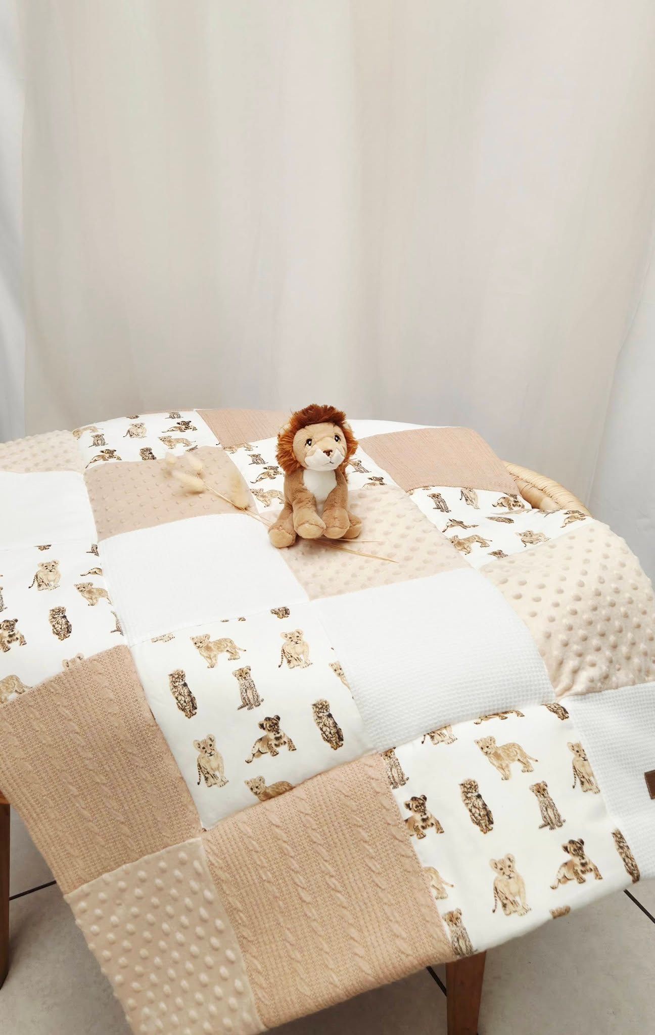 Neutral beige patchwork baby quilt with lion print fabric squares and a small stuffed lion toy displayed on a wooden table.