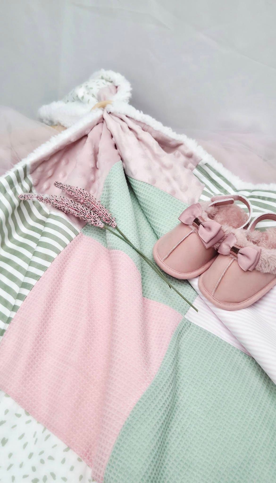 Pink baby shoes with bows rest on a pastel pink and green patchwork blanket with a stuffed bunny toy.