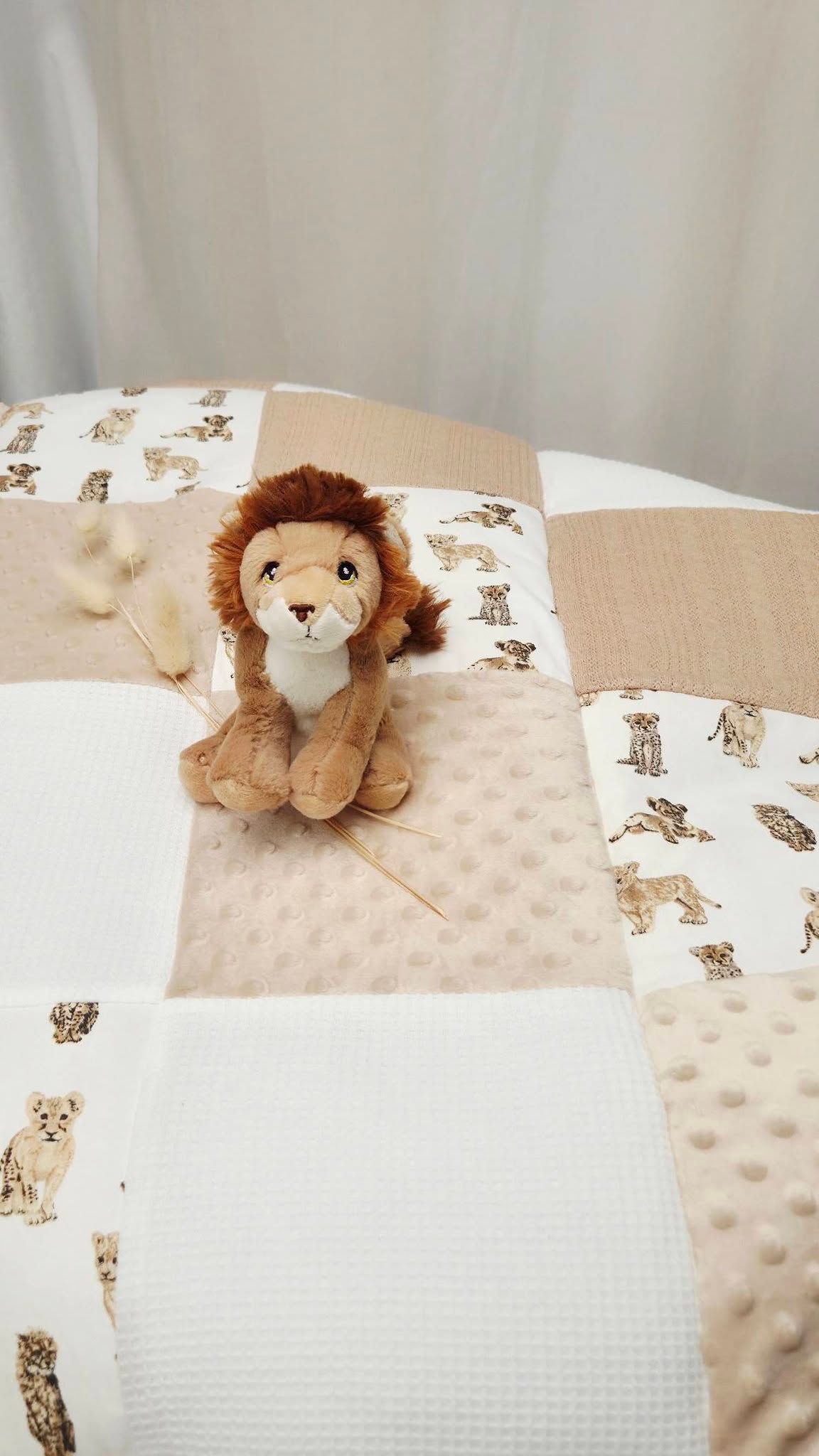 Lion stuffed animal toy sitting on a patchwork baby quilt with safari animal print in neutral beige tones.