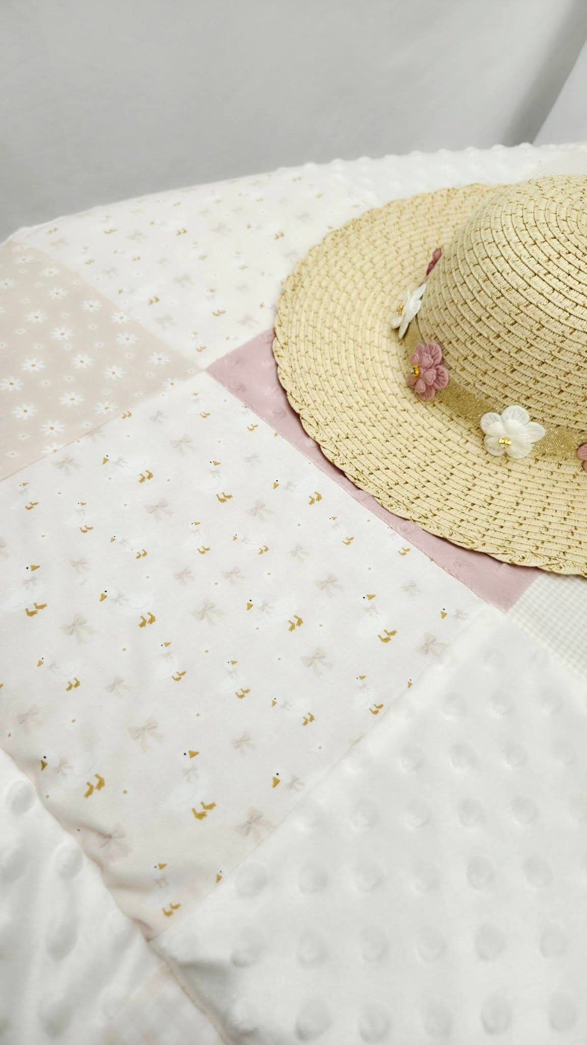 A straw sun hat with pink flowers rests on a white textured blanket with delicate gold pattern details.