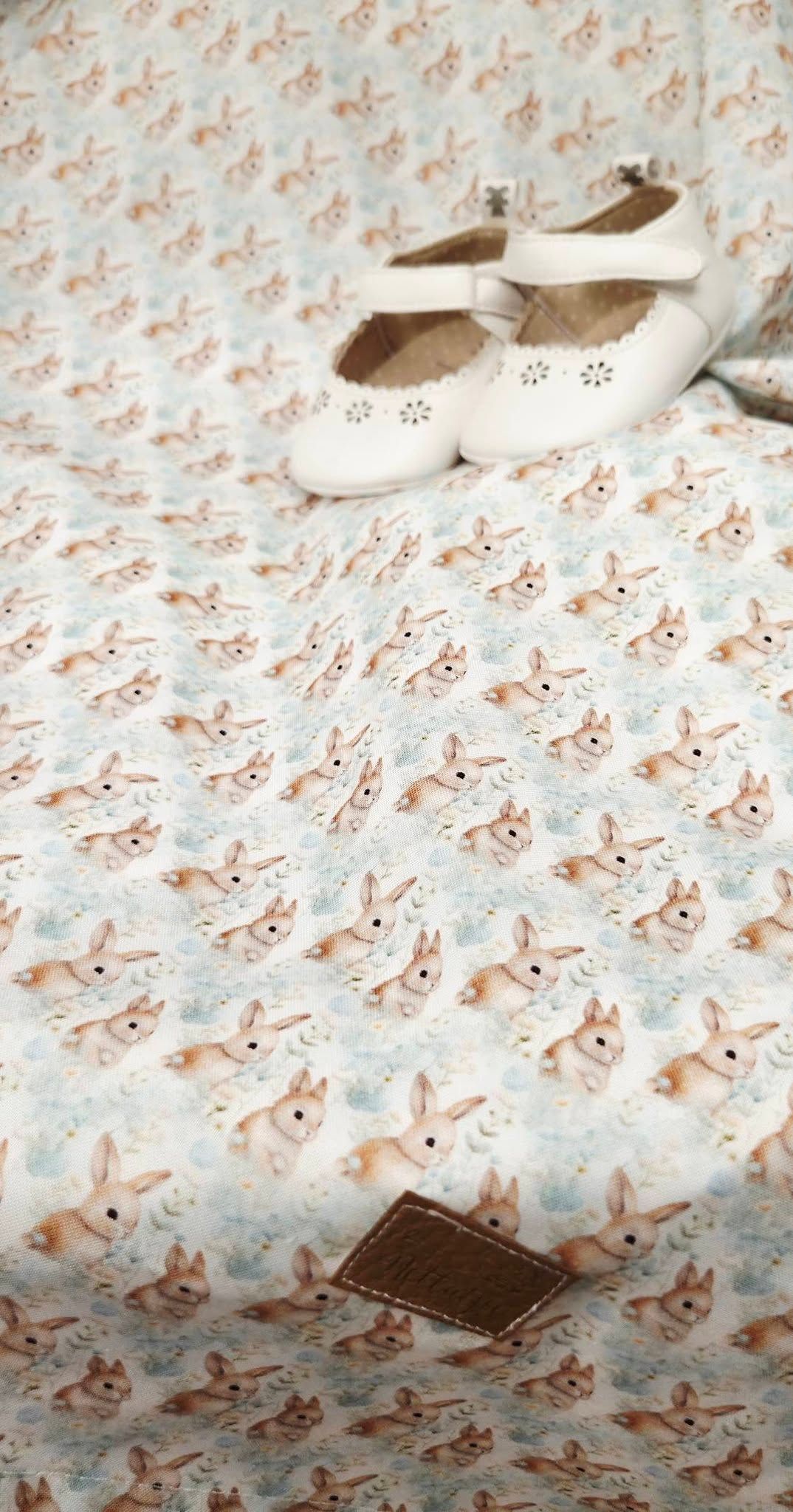 Bunny print fabric spread flat with white Mary Jane shoes and a brown leather tag on top.