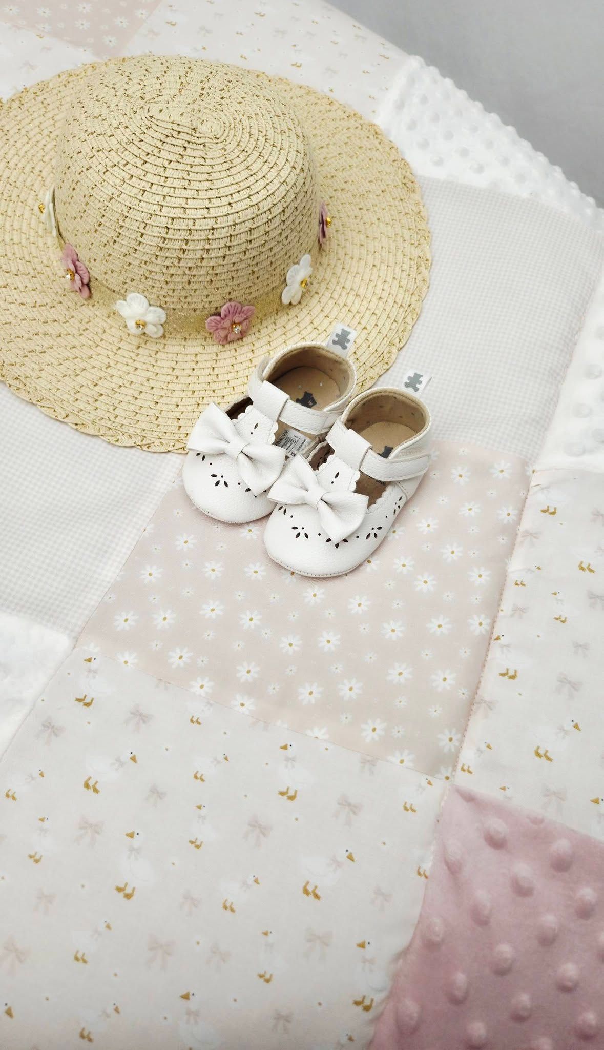 Baby girl straw sun hat with floral details and white bow mary jane shoes on a pastel pink patchwork quilt.