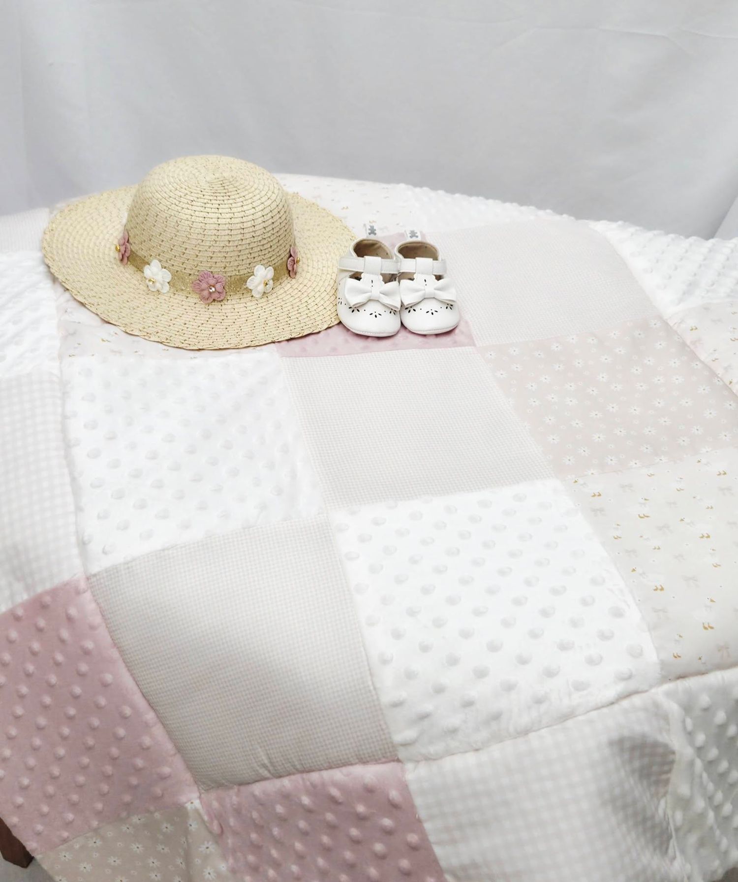 Pink and white patchwork baby quilt with a straw sun hat and white baby shoes displayed on top.