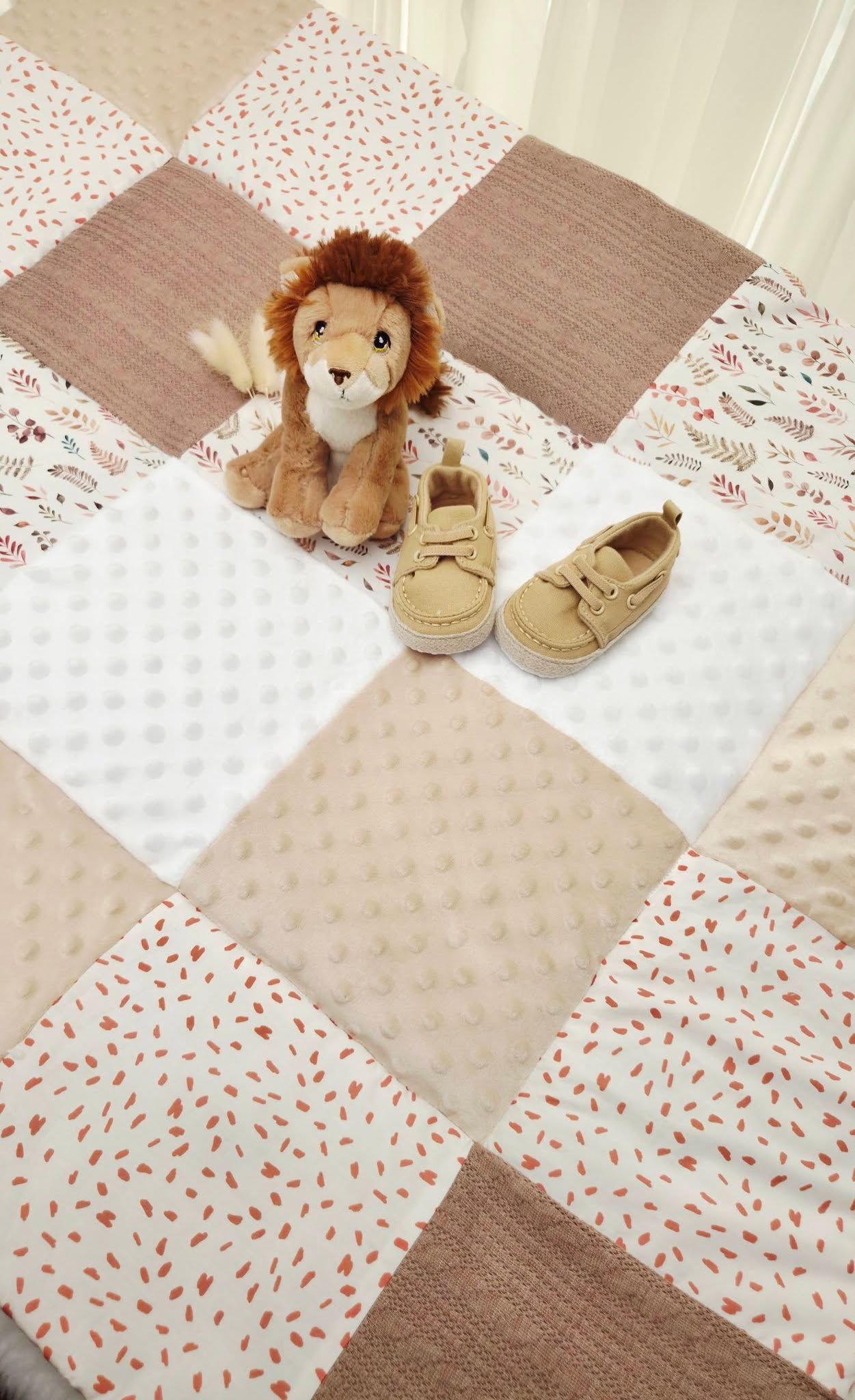 Baby quilt with patchwork design featuring a plush lion stuffed animal and tan infant shoes centerpiece.