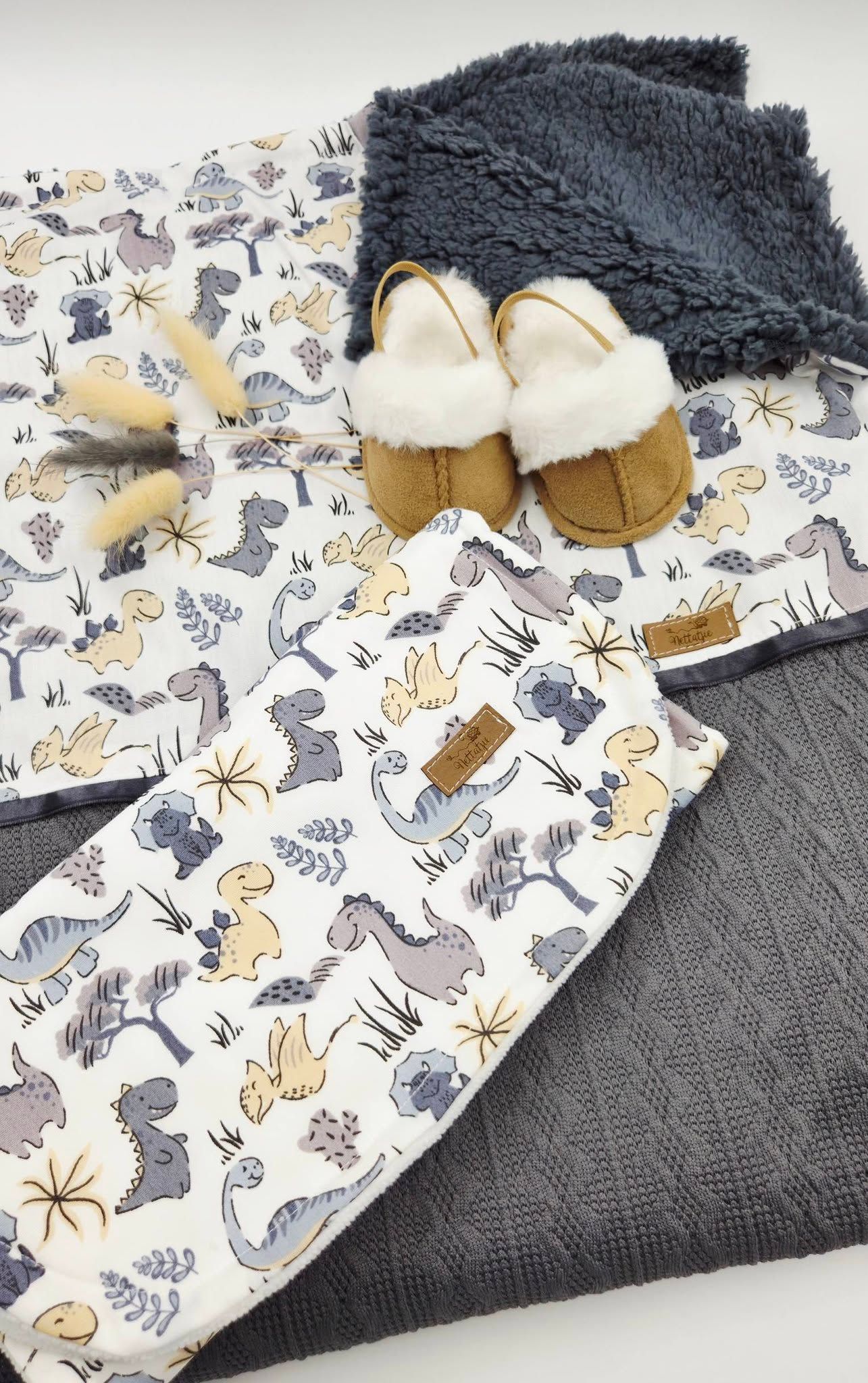 Baby dinosaur print blankets with gray sherpa, knit throw, tan fur slippers, and wooden teether toy flat lay.