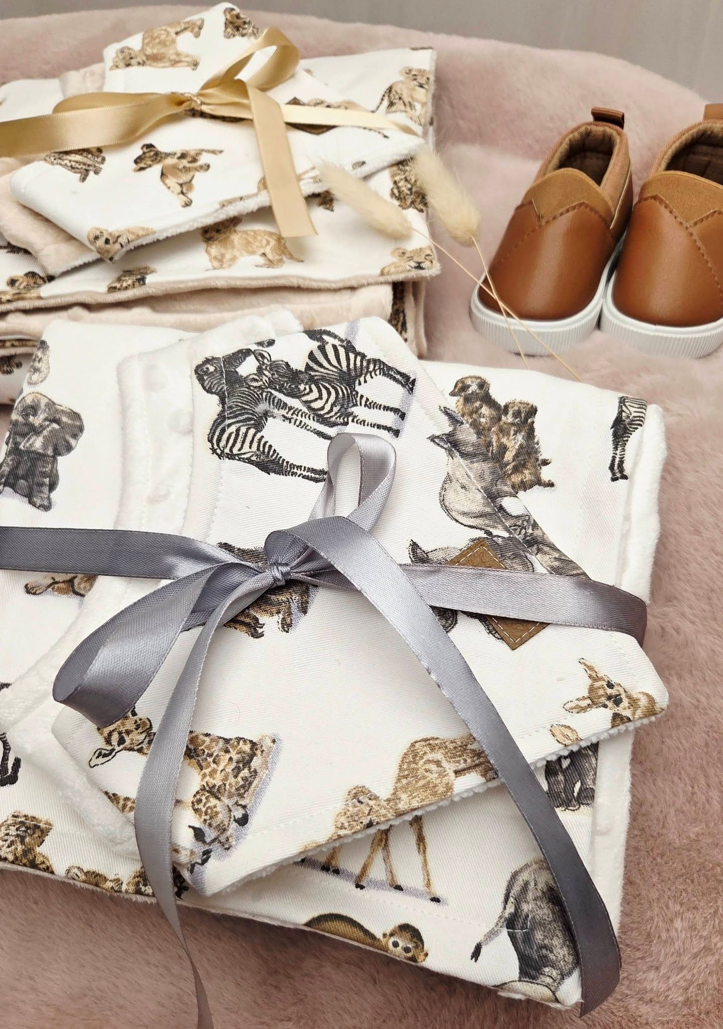 Folded baby blankets with animal print and ribbon bows displayed next to tan leather baby shoes on a pink surface.