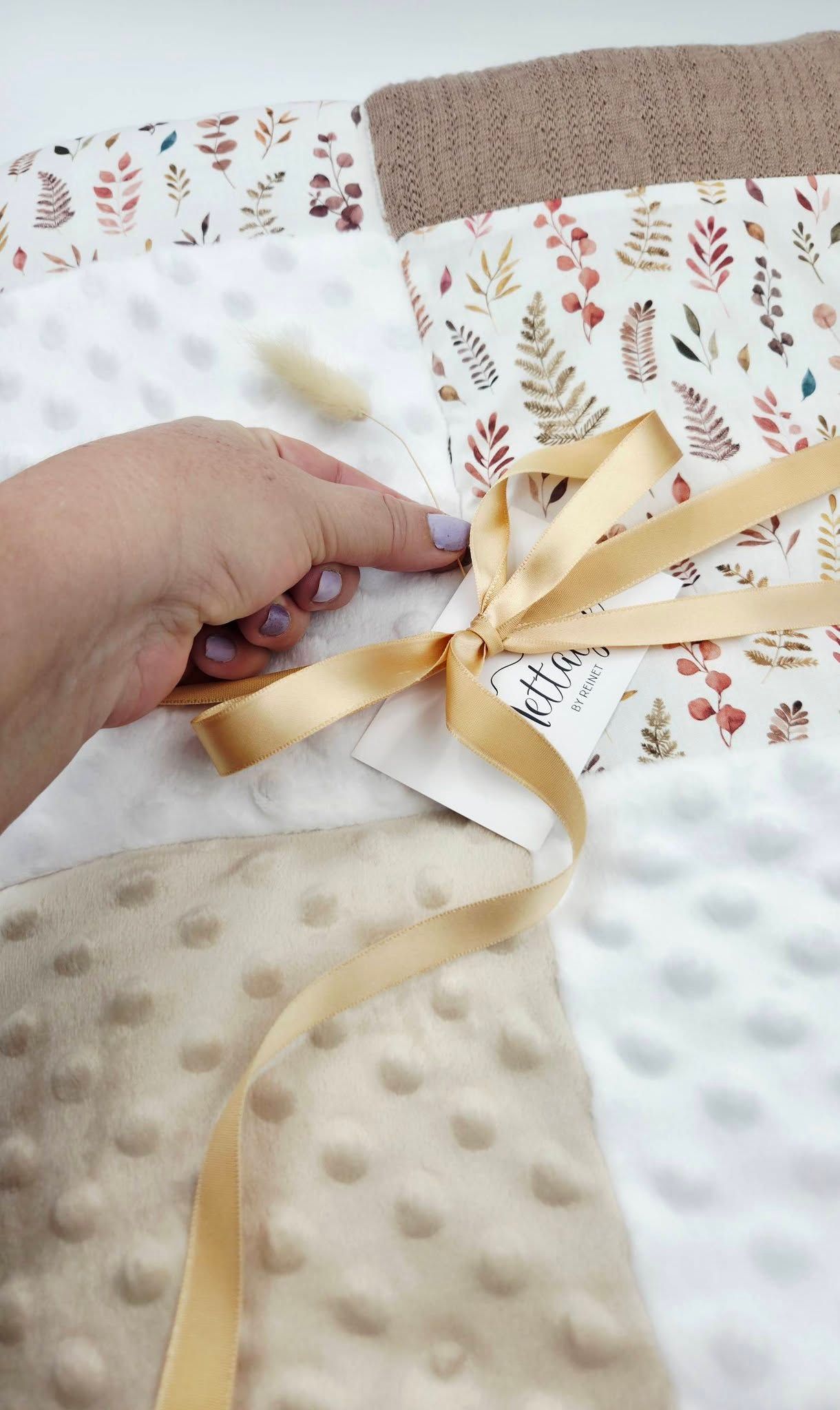 Hand holding a gold ribbon bow on a patchwork baby blanket with floral print and beige minky dot fabric.