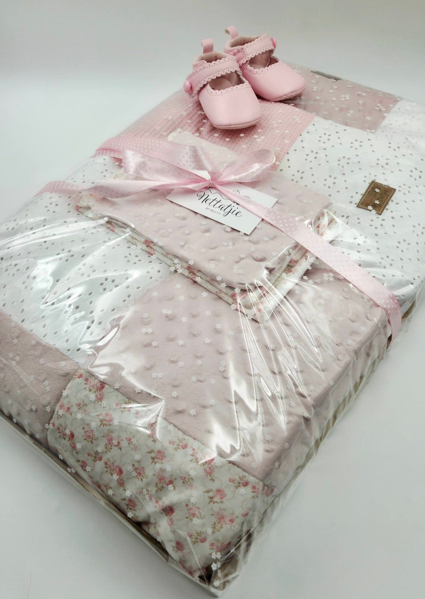Pink baby gift set with floral patchwork blanket, satin ribbon, and tiny pink Mary Jane shoes in clear packaging.