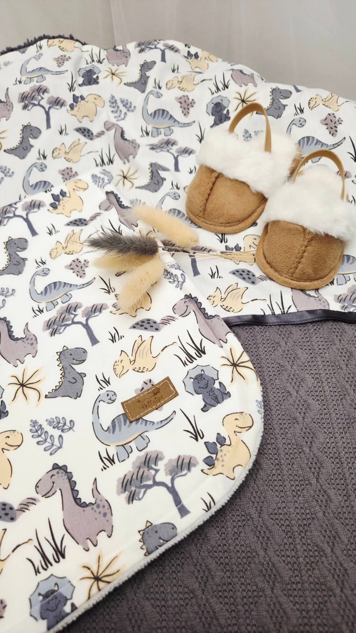 Soft dinosaur print baby blanket with tan fur-trimmed baby slippers on a gray knit background.