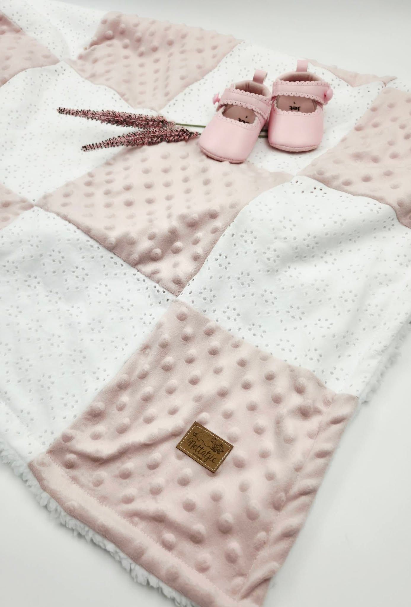 Pink patchwork baby blanket with minky dot texture, paired with pink baby shoes and lavender sprigs on white background.