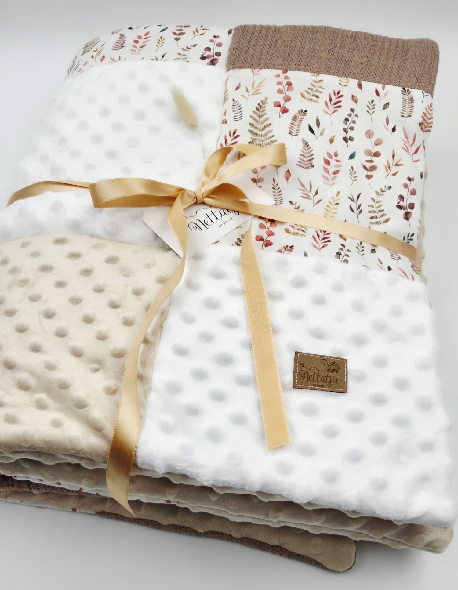 Cream minky dot baby blanket with floral patchwork top, tied with golden satin ribbon, featuring a branded leather tag.