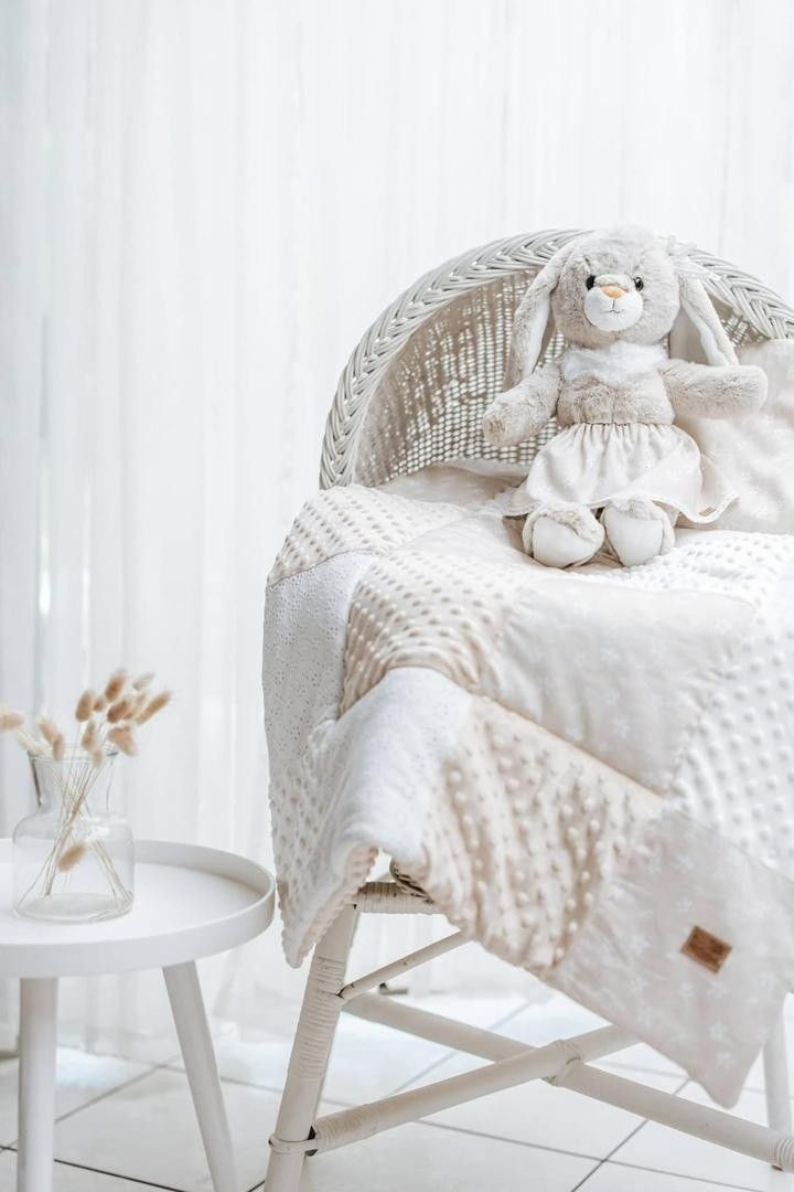 White wicker chair with a soft bunny stuffed animal and white quilted blanket, next to a small table with dried flowers.