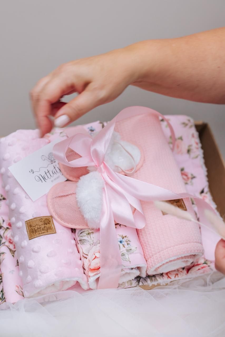 Hands carefully unfold a series of delicate pink baby items with bows and floral patterns on a white surface.