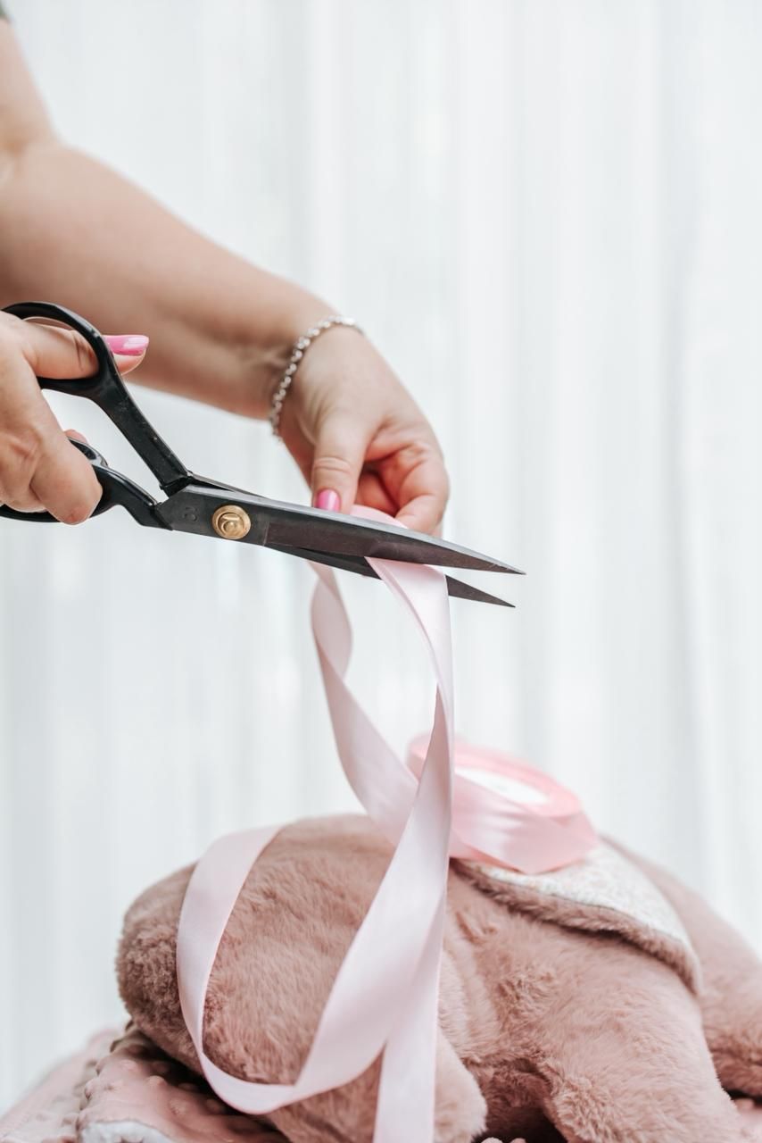 Hands cutting pink ribbon with scissors on plush pink toy for baby announcement.