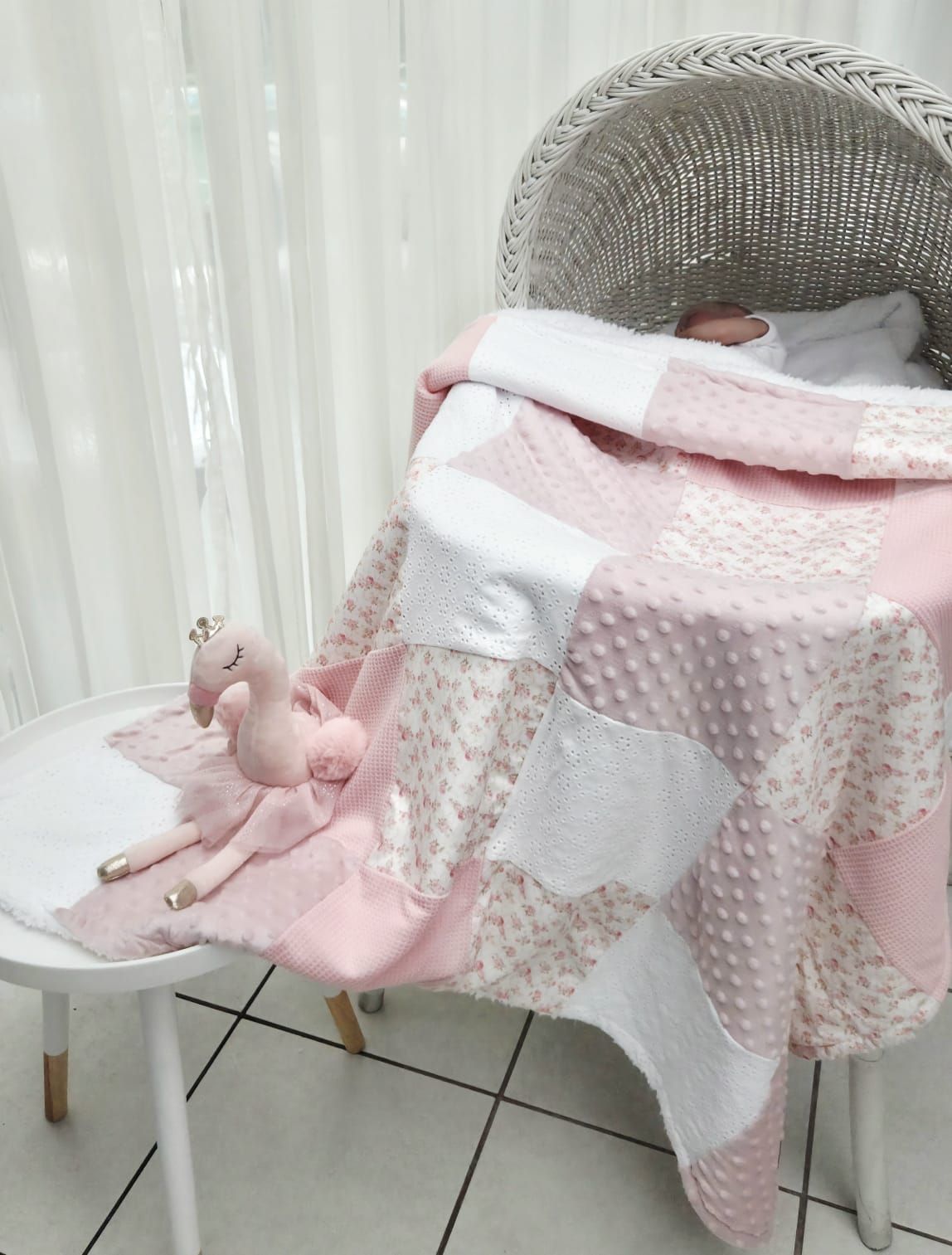 Pink and white patchwork baby blanket draped over a wicker bassinet with a plush flamingo toy on a white chair.